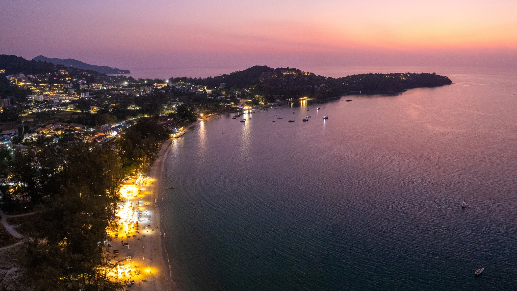 Luftaufnahme einer Küstenstadt bei Sonnenuntergang mit beleuchteten Gebäuden und ruhigem Meer