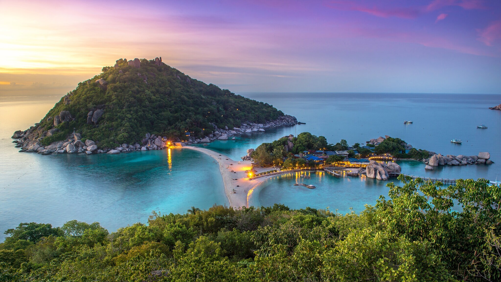 Blick auf eine tropische Insel mit bewaldetem Hügel, Sandbank und beleuchteten Gebäuden bei Sonnenuntergang