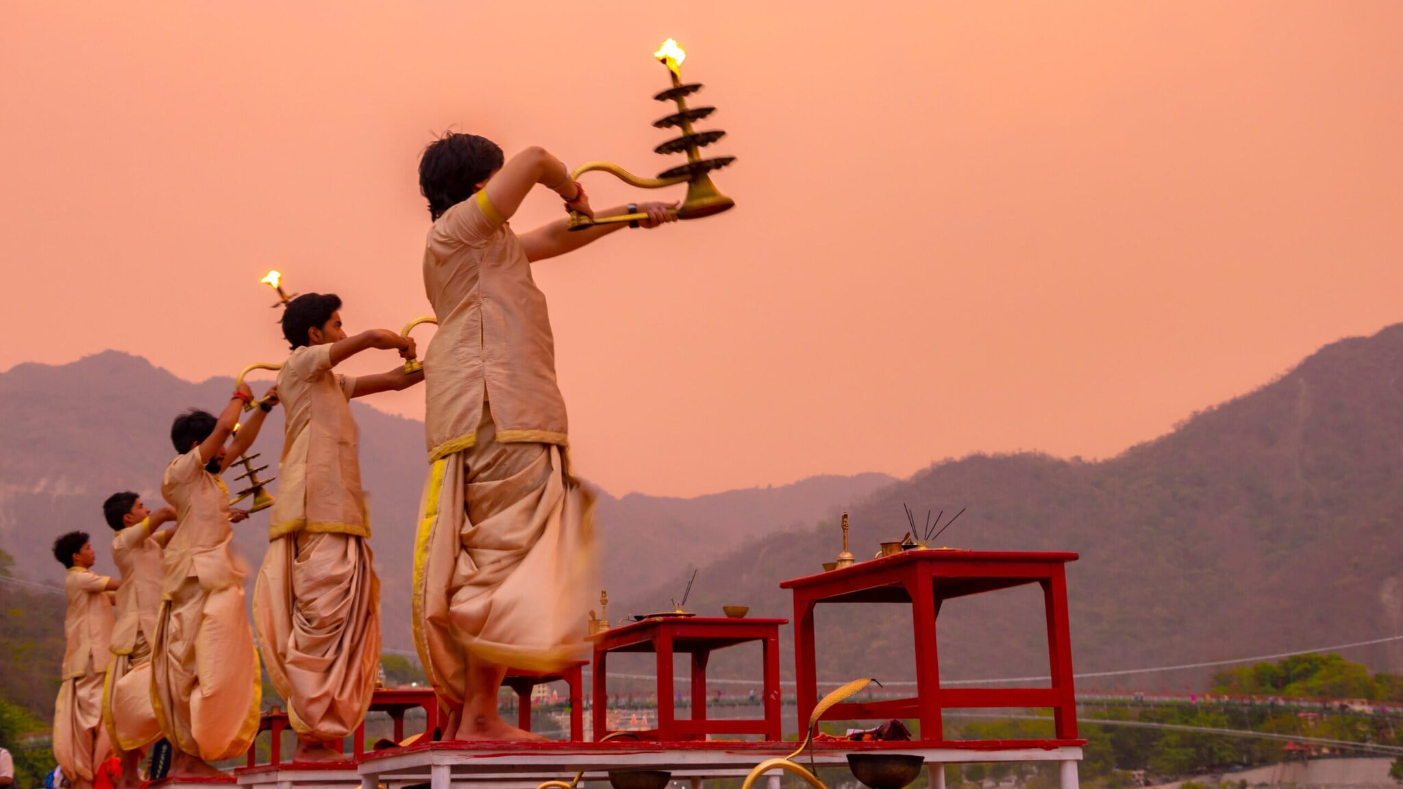 Fünf Personen in traditioneller Kleidung führen eine rituelle Zeremonie mit brennenden Öllampen am Flussufer bei Sonnenuntergang durch Fünf Personen in traditioneller Kleidung führen eine rituelle Zeremonie mit brennenden Öllampen am Flussufer bei Sonnenuntergang durch