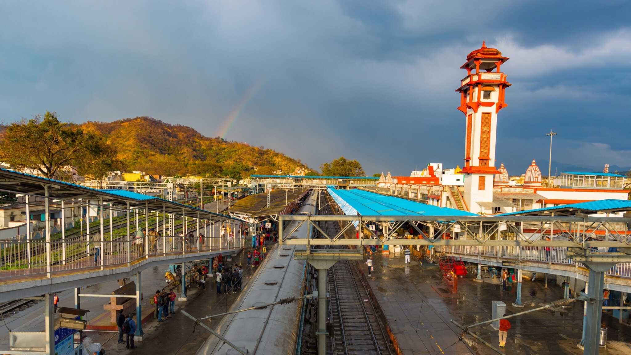 Blick auf einen Bahnhof mit Bahnsteigen und einem Zug, im Hintergrund ein hoher Turm und bewaldete Hügel unter bewölktem Himmel Blick auf einen Bahnhof mit Bahnsteigen und einem Zug, im Hintergrund ein hoher Turm und bewaldete Hügel unter bewölktem Himmel