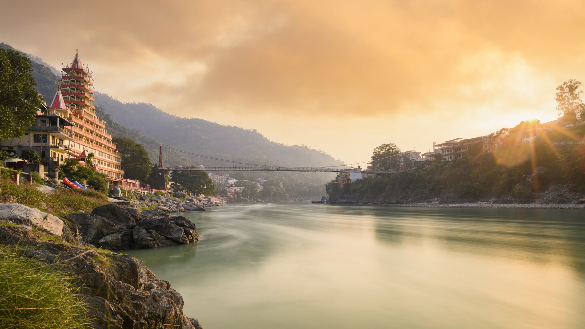 Fluss mit hängender Fußgängerbrücke und mehrstöckigem Tempelgebäude am Ufer bei Sonnenuntergang in bergiger Landschaft Fluss mit hängender Fußgängerbrücke und mehrstöckigem Tempelgebäude am Ufer bei Sonnenuntergang in bergiger Landschaft