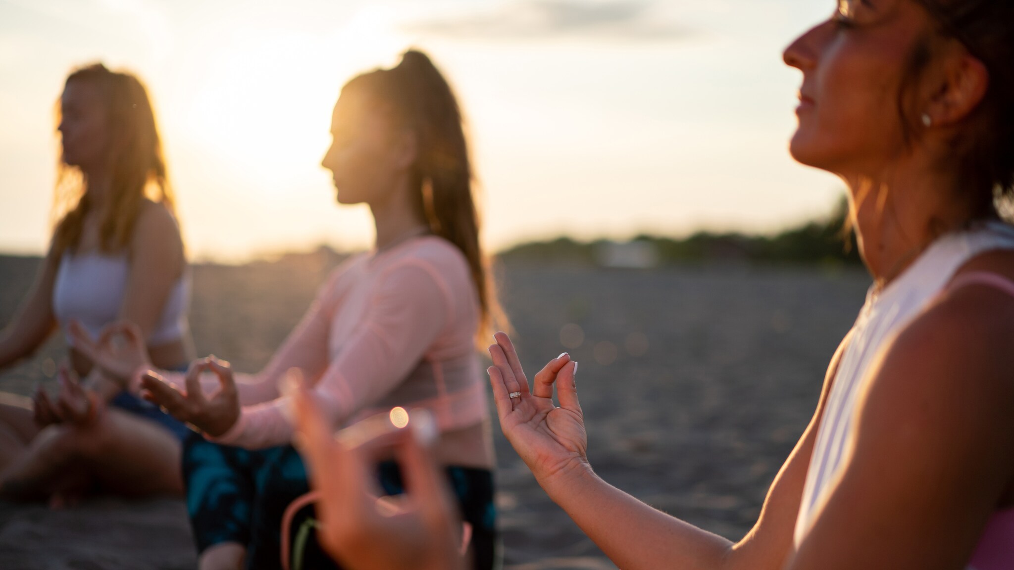 Drei Frauen sitzen im Schneidersitz am Strand und meditieren bei Sonnenuntergang