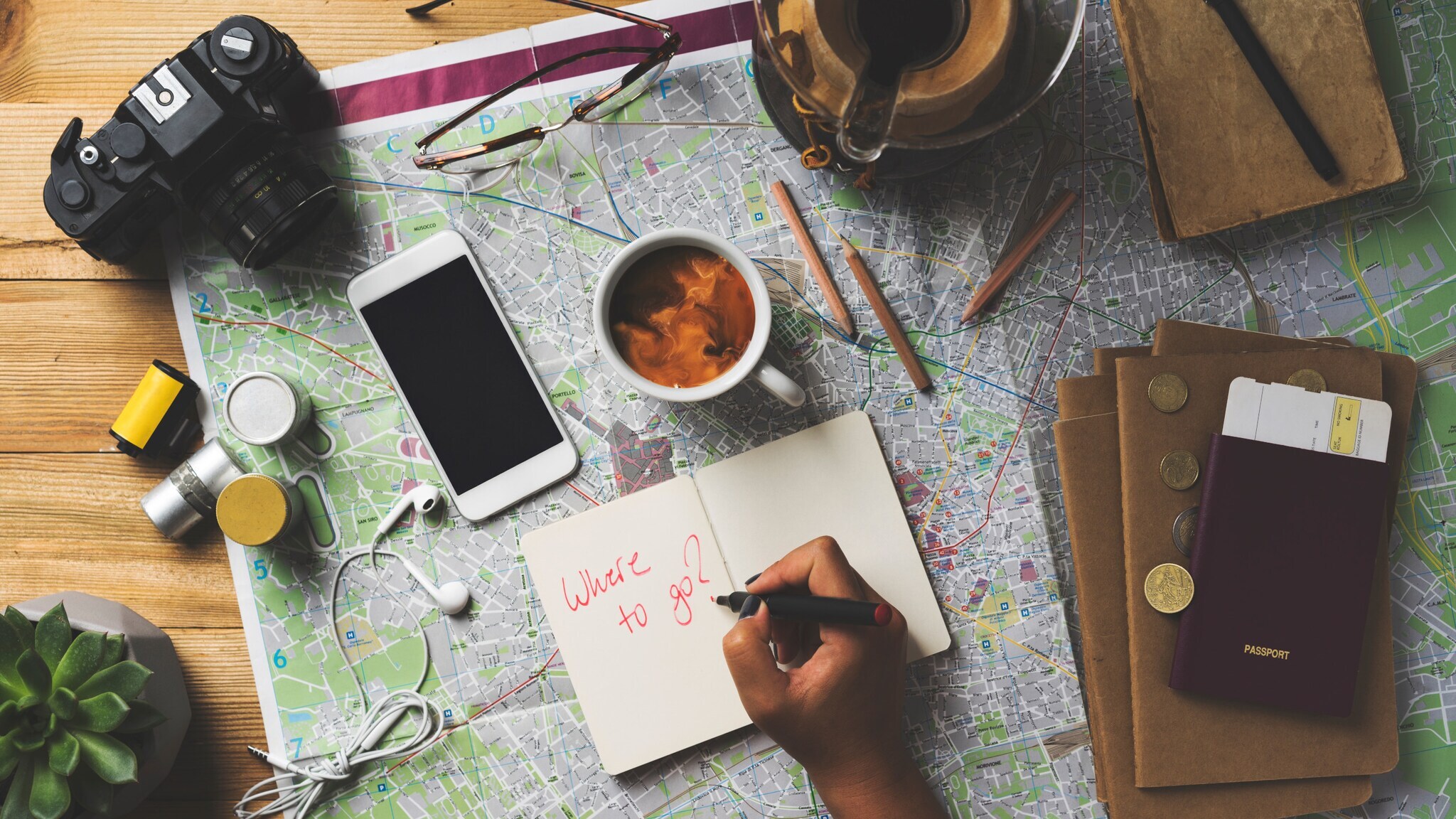 Hand schreibt 'Where to go?' in Notizbuch auf Landkarte mit Kamera, Smartphone, Kaffeetasse, Brille, Reisepass und Münzen