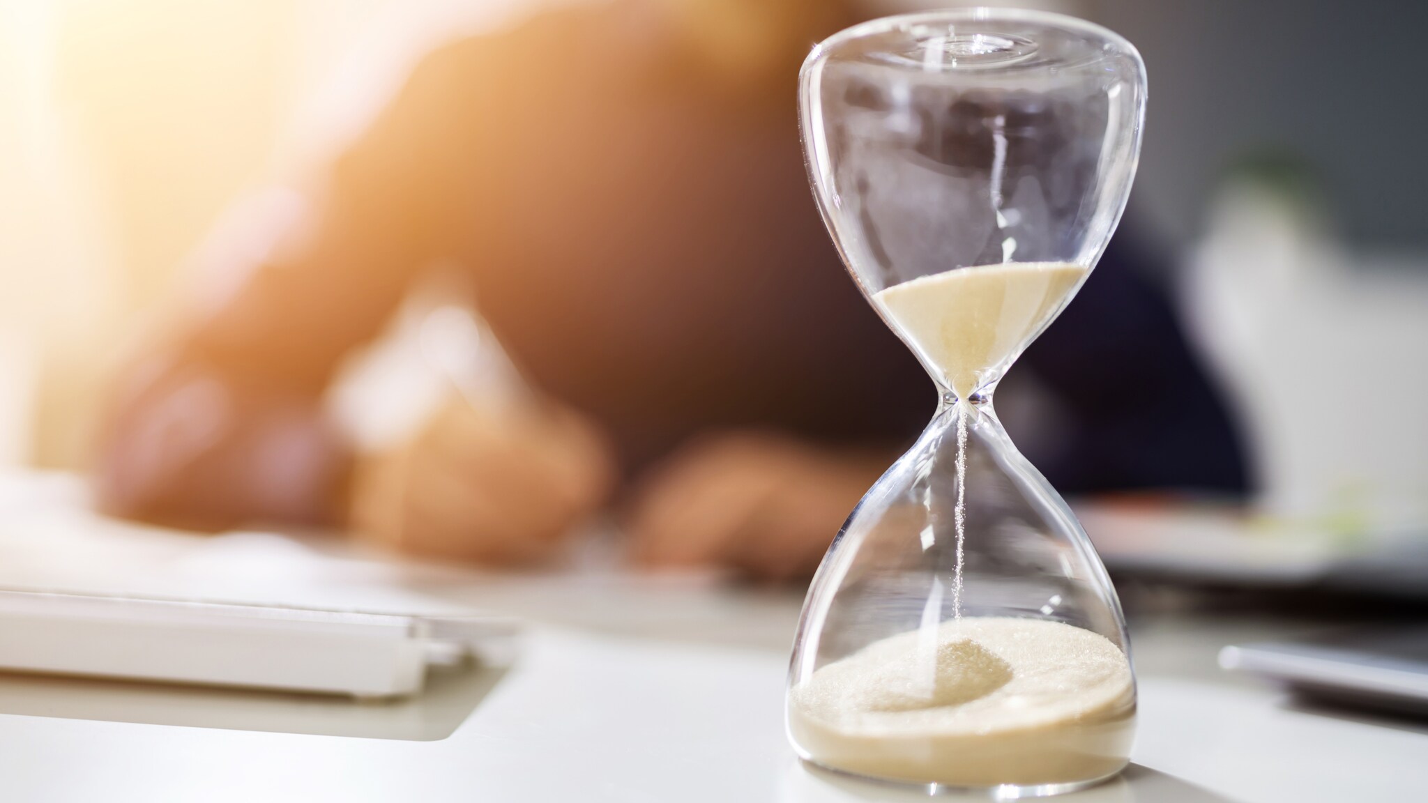 Sanduhr auf einem Schreibtisch mit verschwommenem Hintergrund einer Person, die den Kopf auf den Tisch legt Sanduhr auf einem Schreibtisch mit verschwommenem Hintergrund einer Person, die den Kopf auf den Tisch legt