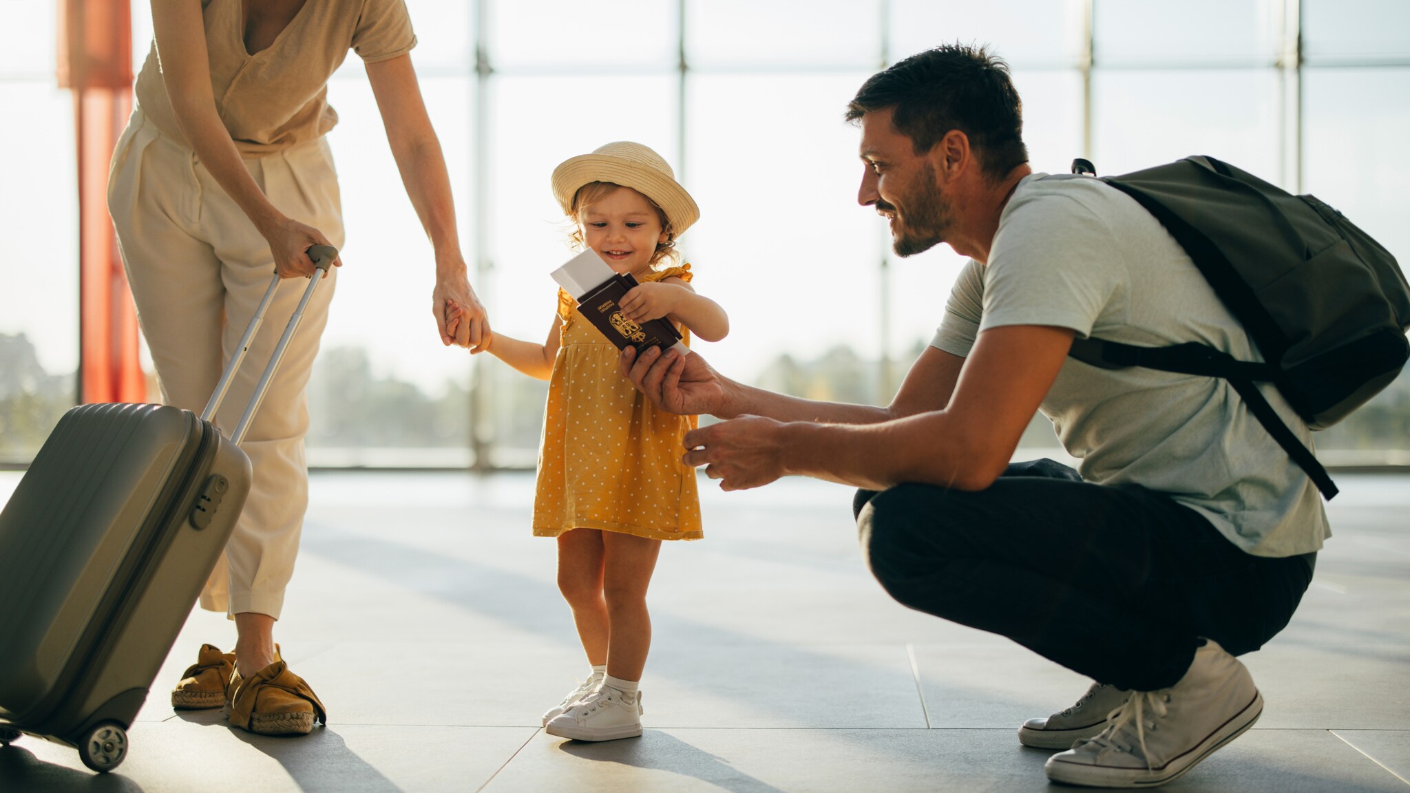 Mutter mit Rollkoffer hält die Hand eines kleinen Mädchens in gelbem Kleid und Sonnenhut, Vater kniet und zeigt Reisepässe Mutter mit Rollkoffer hält die Hand eines kleinen Mädchens in gelbem Kleid und Sonnenhut, Vater kniet und zeigt Reisepässe