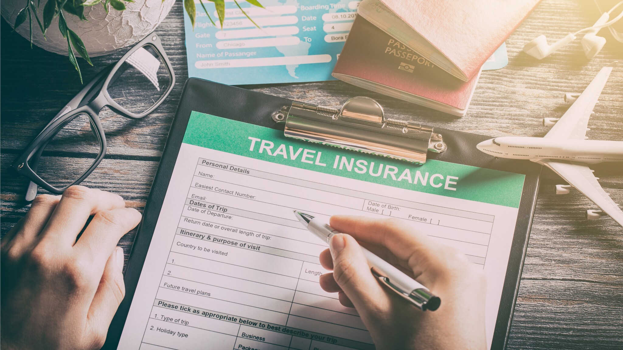 Person füllt ein Formular zur Reiseversicherung auf einem Klemmbrett aus, umgeben von Reisepässen, Flugtickets, einer Brille und einem Modellflugzeug Person füllt ein Formular zur Reiseversicherung auf einem Klemmbrett aus, umgeben von Reisepässen, Flugtickets, einer Brille und einem Modellflugzeug
