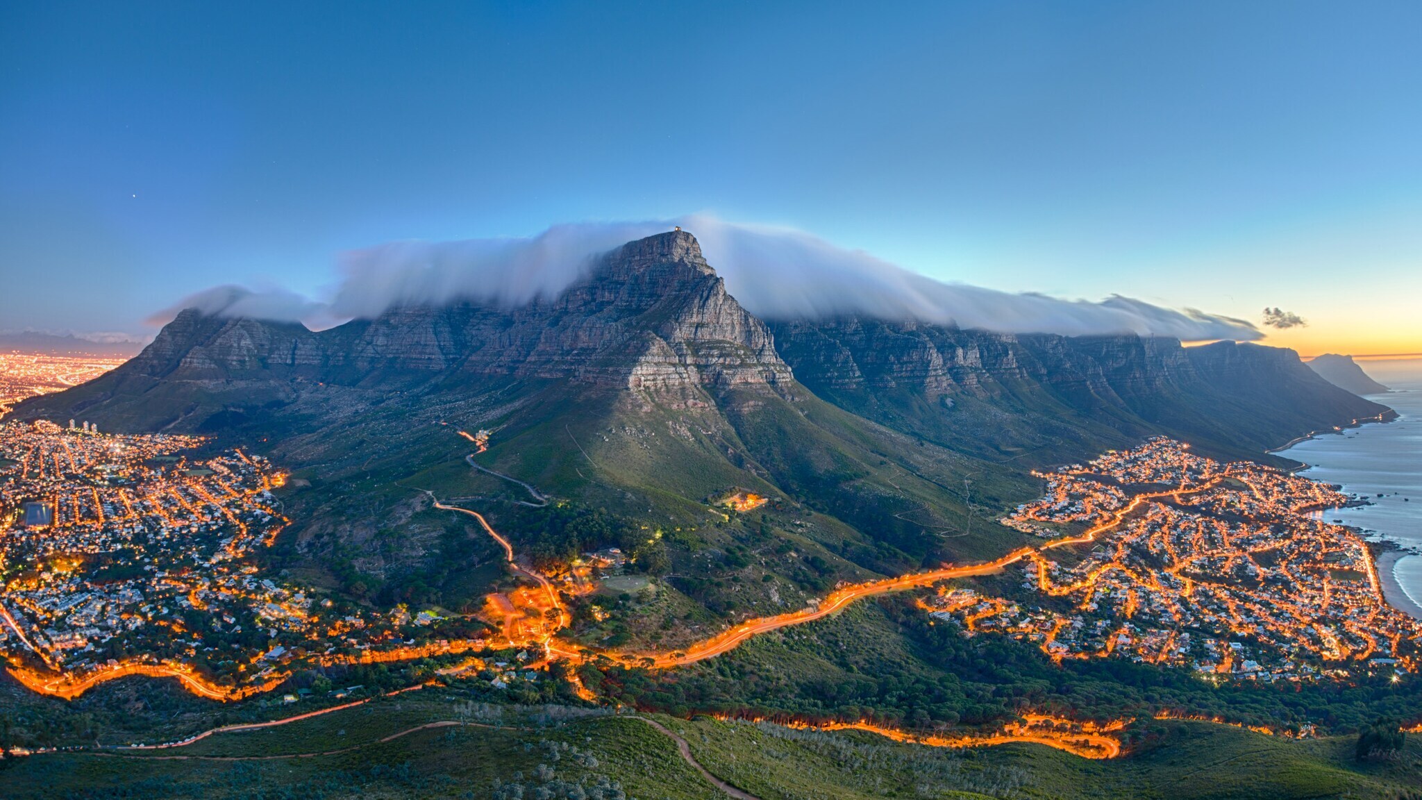 Tafelberg mit Wolkenkappe über Kapstadt bei Sonnenuntergang, beleuchtete Stadt und Küste im Hintergrund