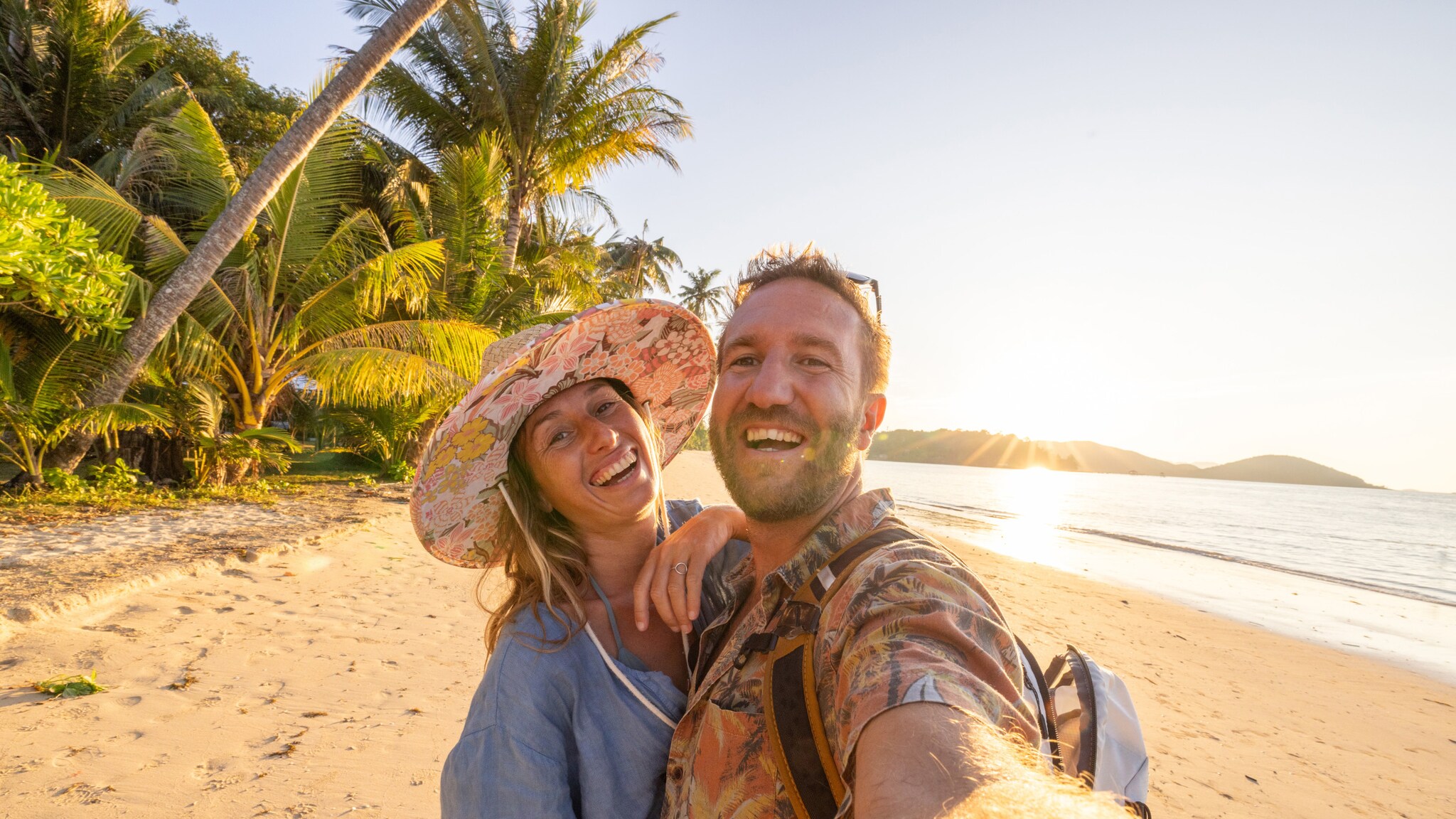 Lachendes Paar macht Selfie am Strand bei Sonnenuntergang, umgeben von Palmen und Sand
