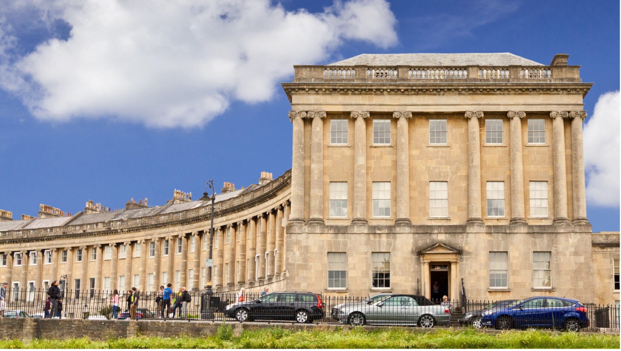 Historisches Gebäude mit Säulen und vielen Fenstern, davor parkende Autos und Menschen auf dem Bürgersteig