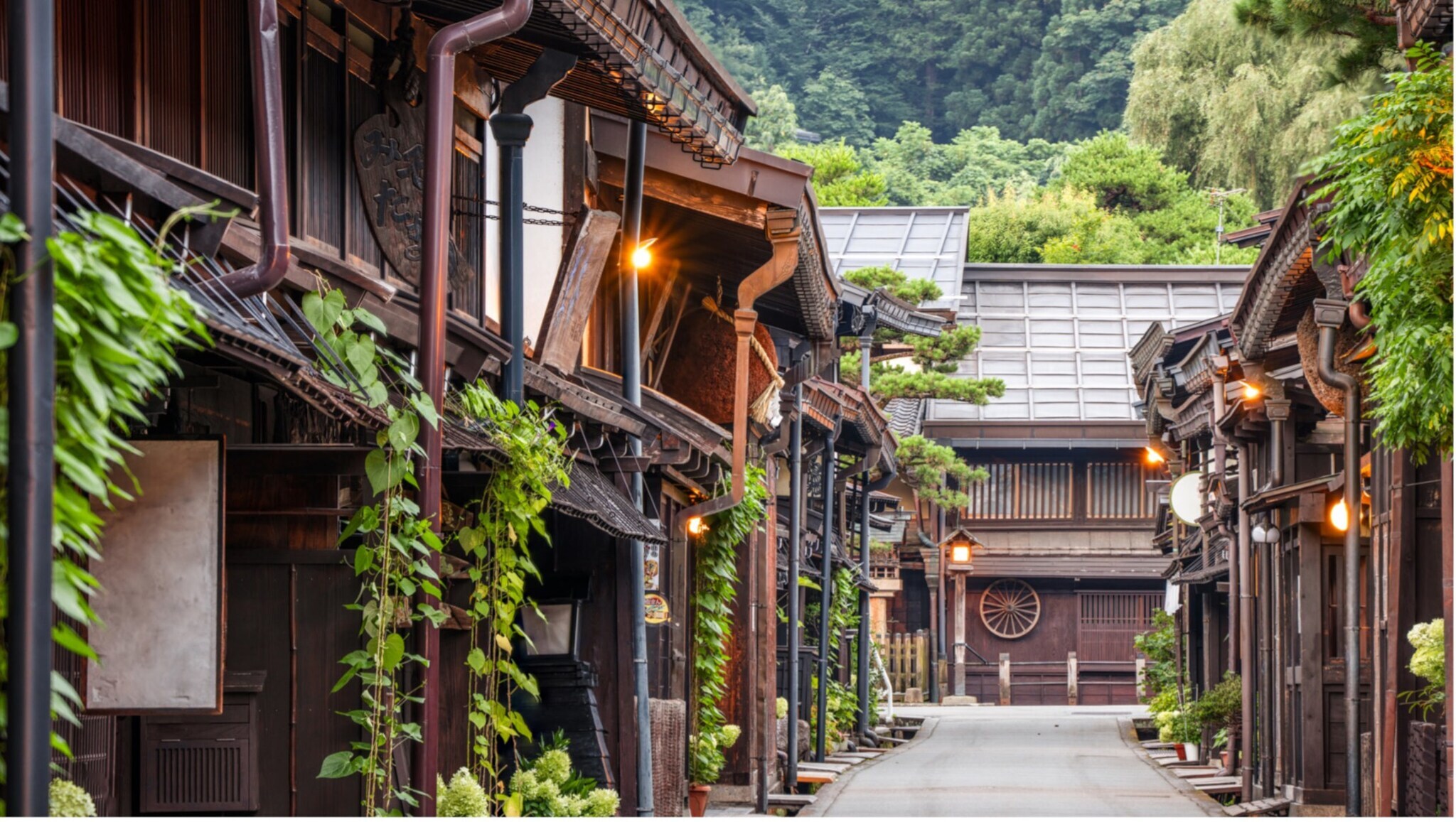 Traditionelle japanische Holzhäuser entlang einer ruhigen Straße mit grünen Pflanzen und bewaldeten Hügeln im Hintergrund