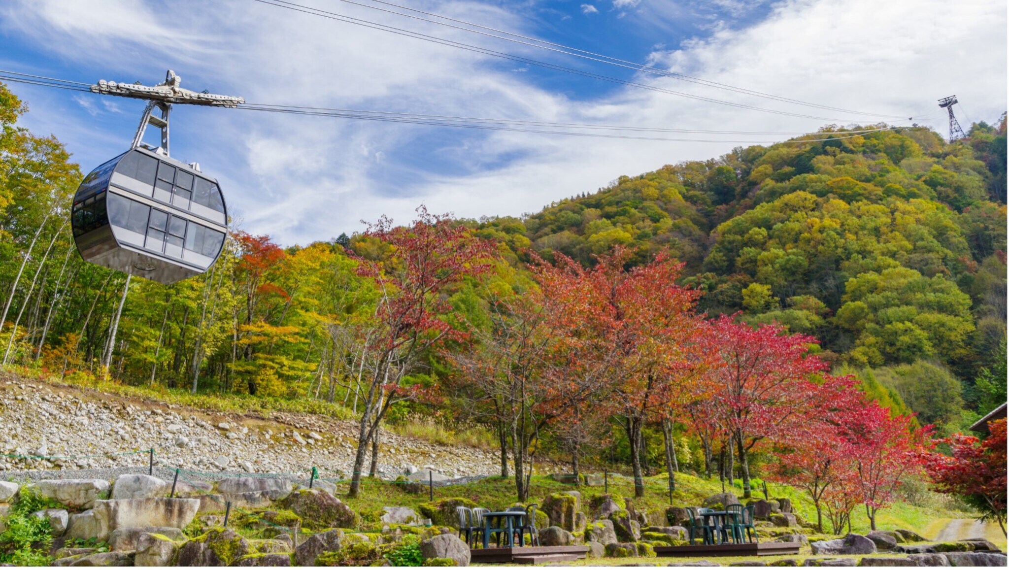 Seilbahn fährt über herbstlich gefärbte Bäume in einem bergigen Landschaftspark mit Sitzbänken und Tischen