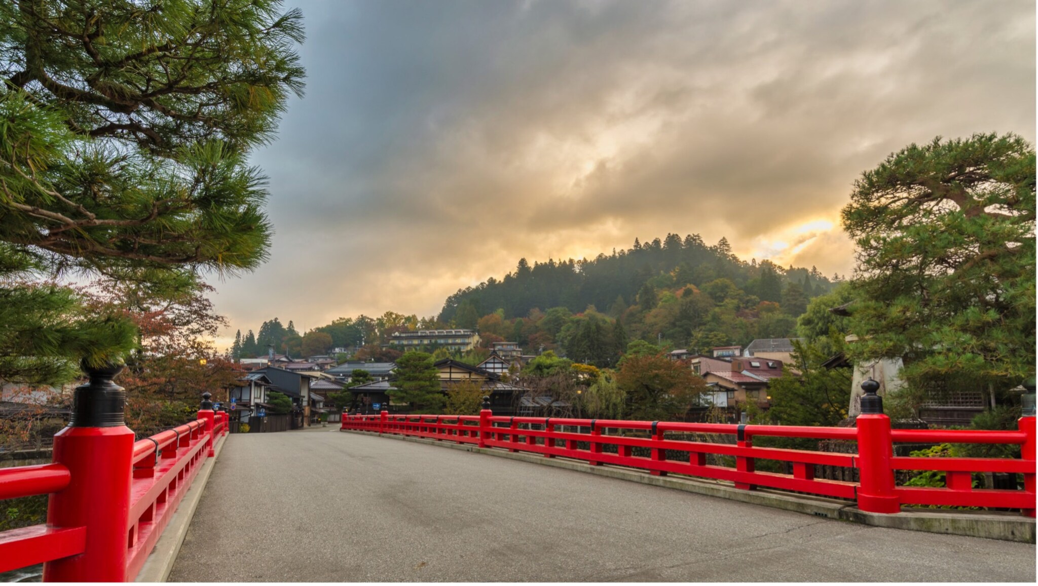 Rote Brücke über Fluss mit Häusern und bewaldetem Hügel im Hintergrund bei bewölktem Himmel Rote Brücke über Fluss mit Häusern und bewaldetem Hügel im Hintergrund bei bewölktem Himmel