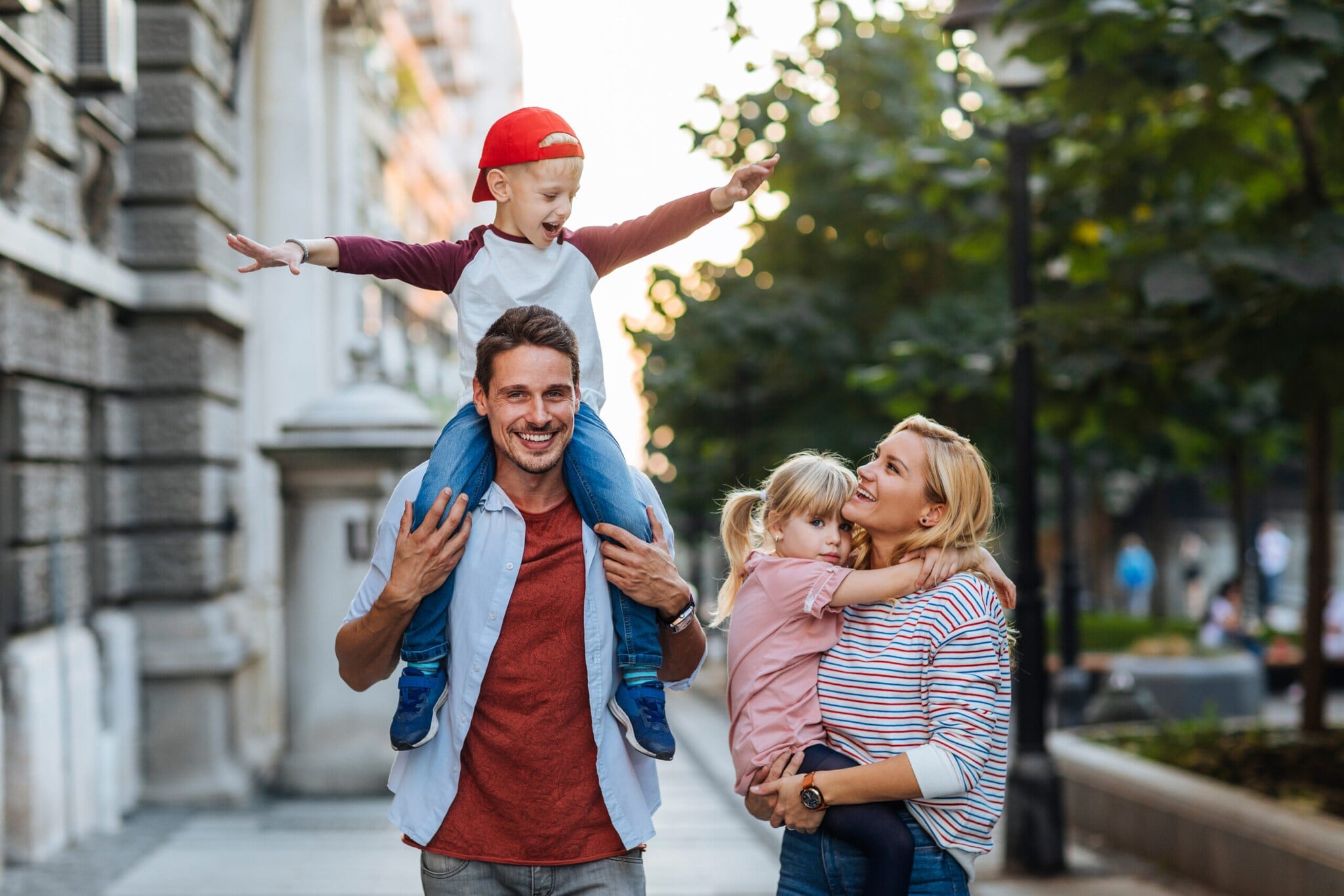 Ein Mann auf einem Weg zwischen Haus und Straße trägt einen Jungen auf den Schultern, eine Frau daneben trägt ein Mädchen. Ein Mann auf einem Weg zwischen Haus und Straße trägt einen Jungen auf den Schultern, eine Frau daneben trägt ein Mädchen.