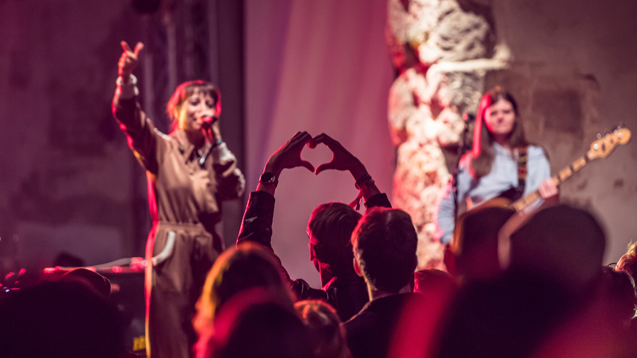 Publikum bei einem Konzert formt mit den Händen ein Herz, während zwei Musikerinnen auf der Bühne spielen