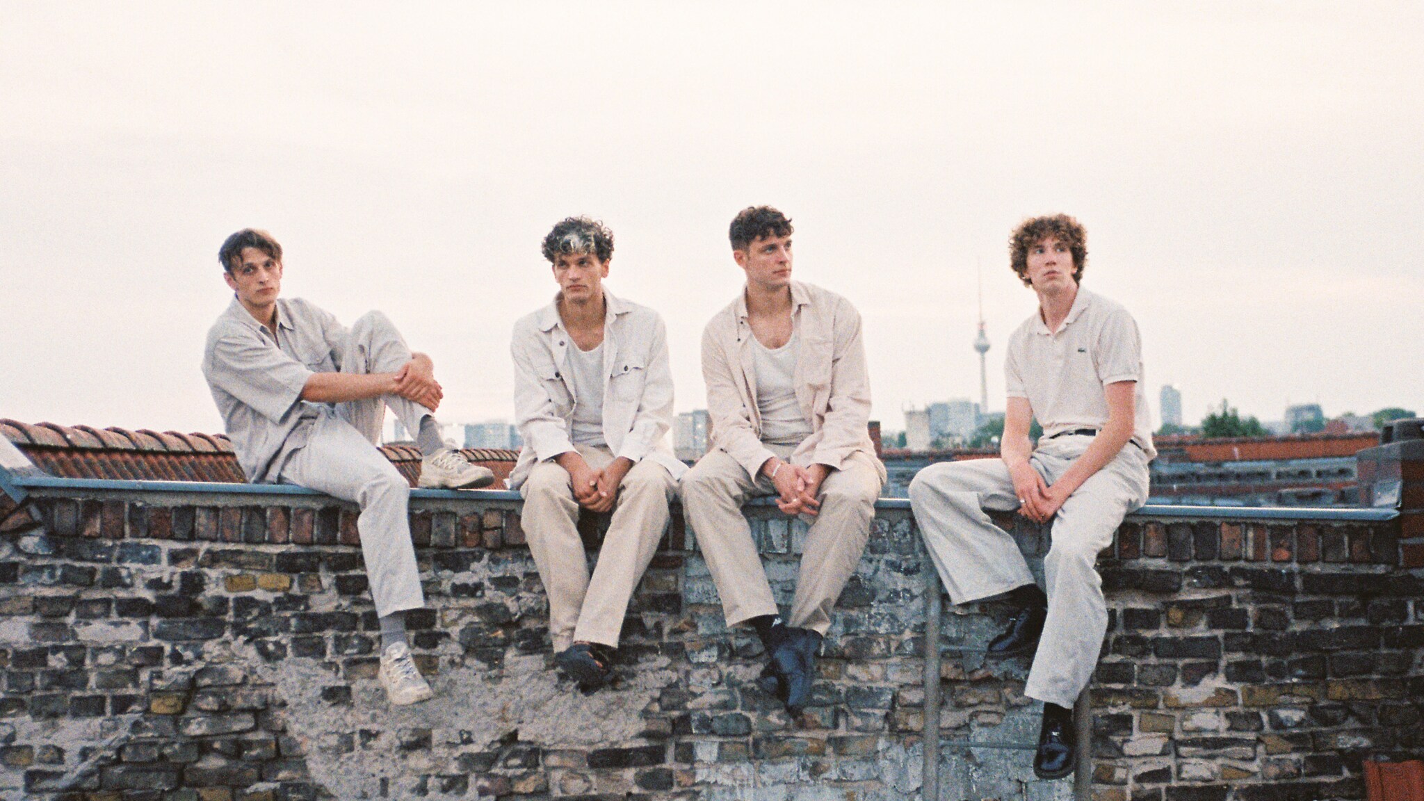 Vier junge Männer, die Mitflieder der Band Oswald, in heller Freizeitkleidung sitzen auf einer Mauer mit Stadtblick im Hintergrund.