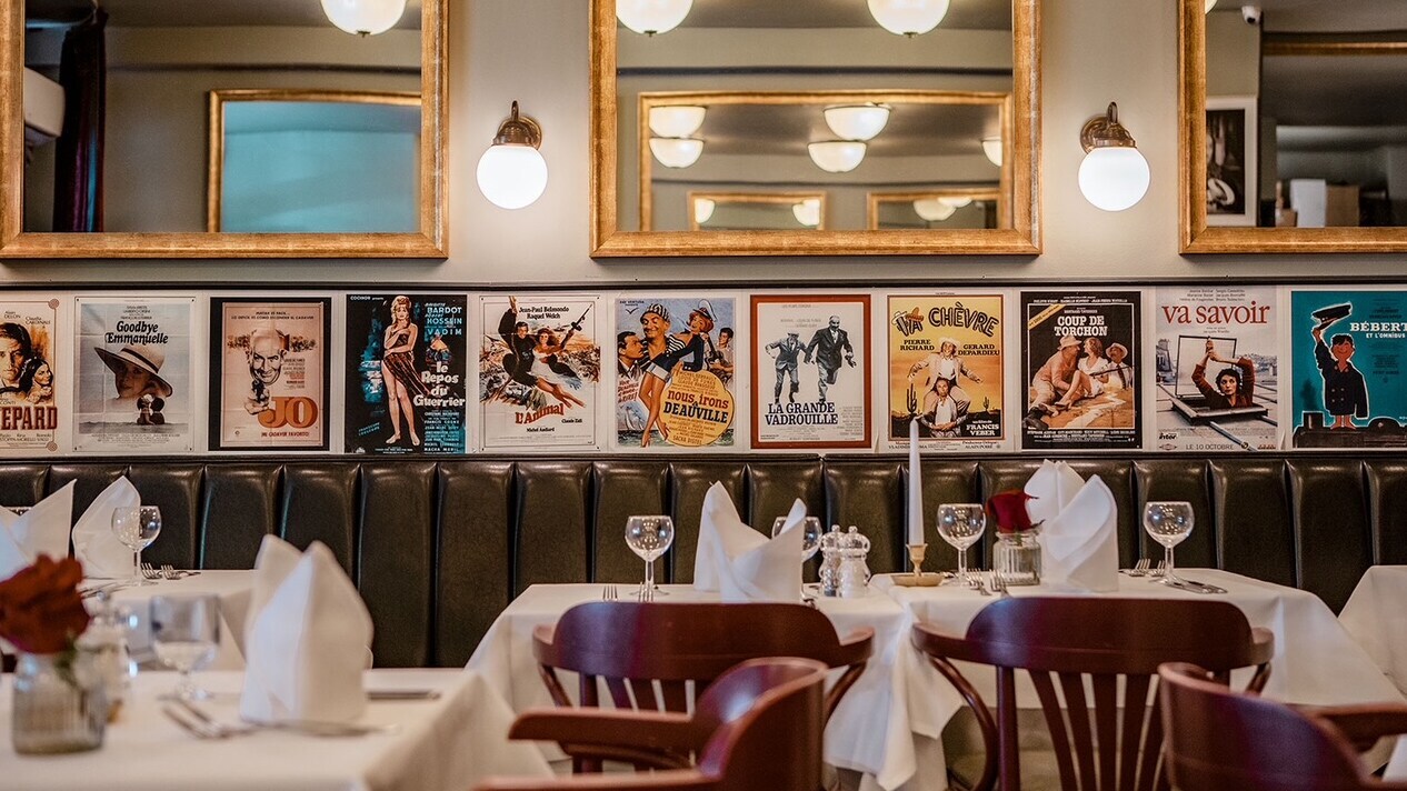 Restaurant mit weißen Tischdecken, gefalteten Servietten, Weingläsern und Stühlen vor einer Wand mit goldgerahmten Spiegeln und Vintage-Filmplakaten