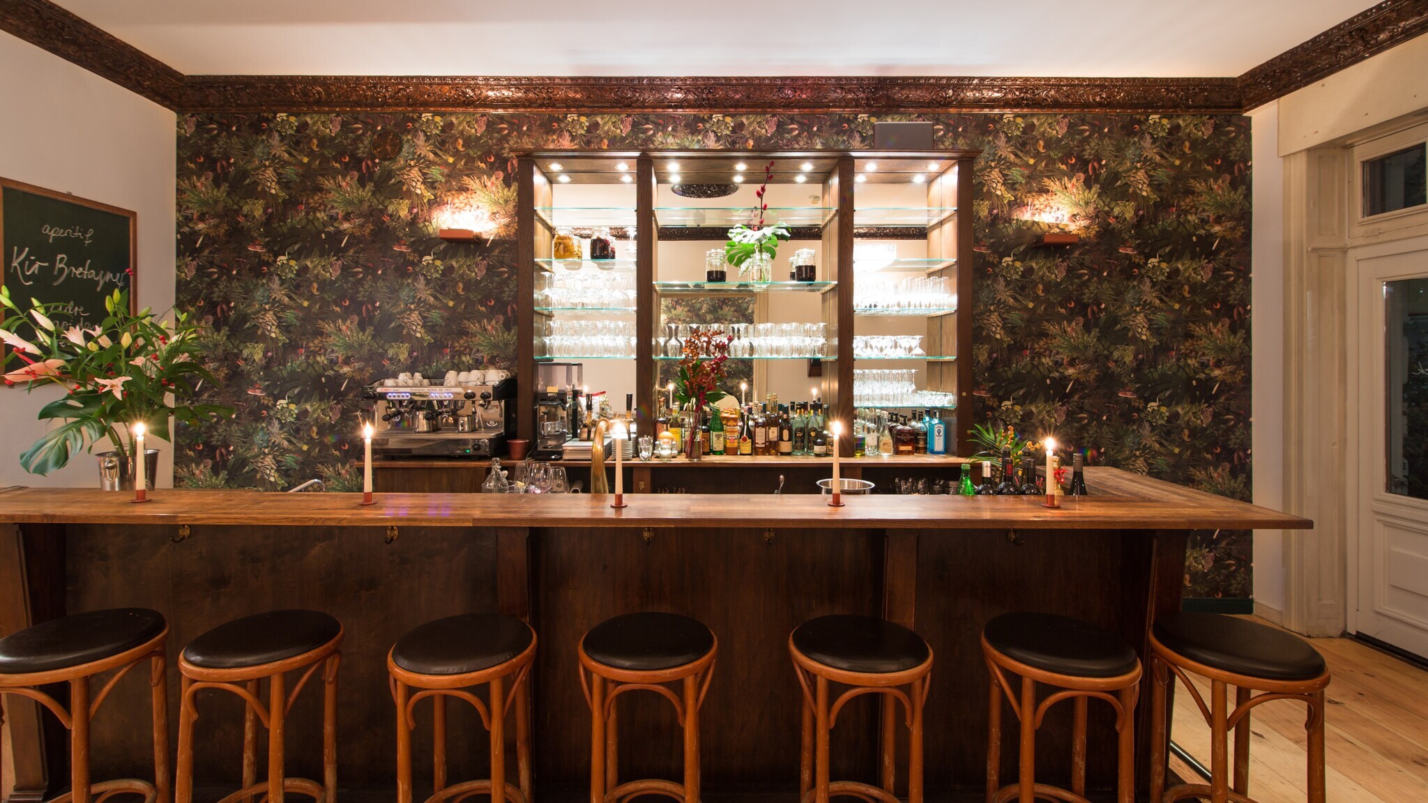 Holztresen mit sieben Barhockern vor einer Wand mit floralem Muster und beleuchtetem Regal mit Gläsern und Flaschen
