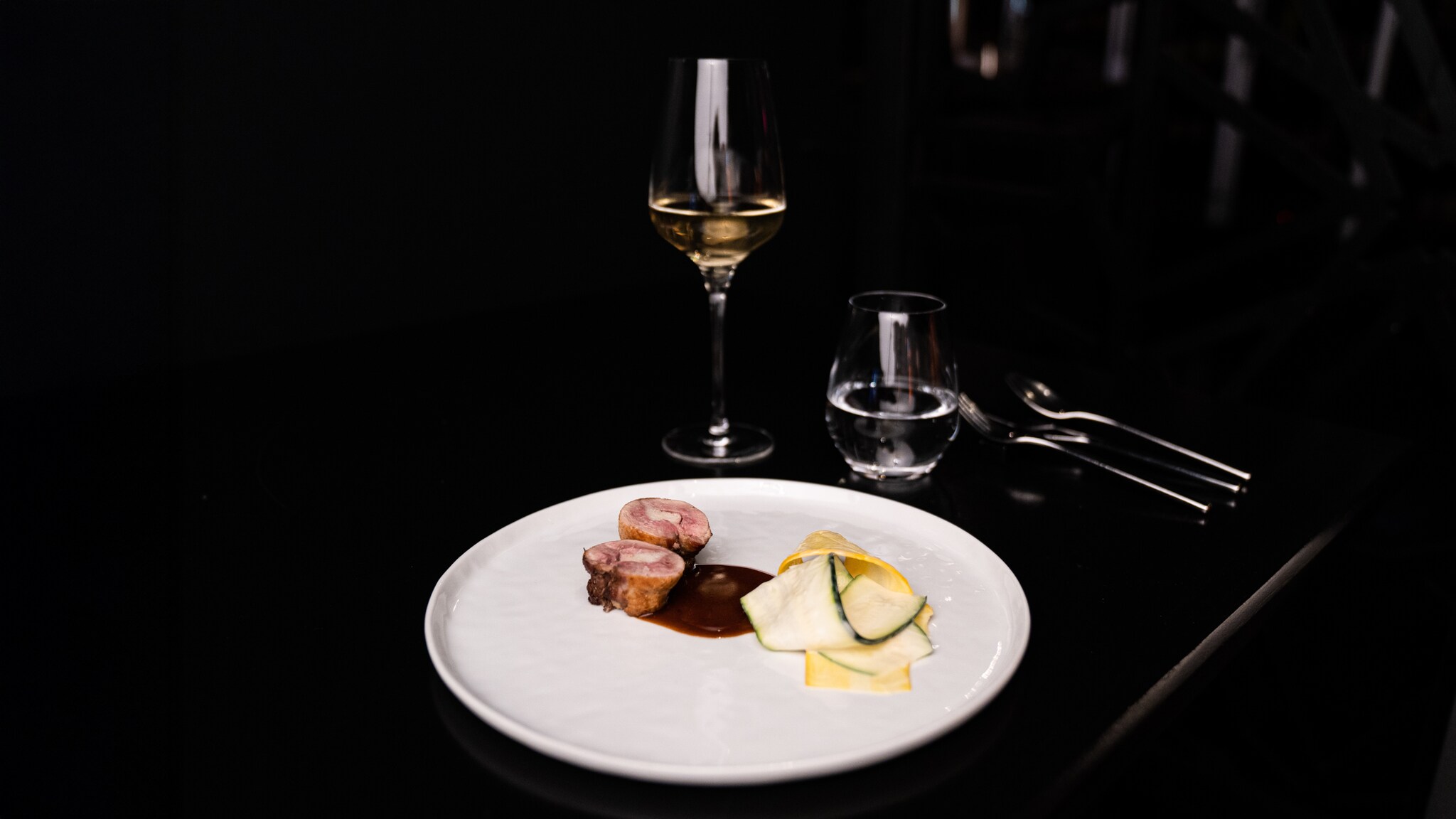 Zwei Scheiben rosa gebratenes Fleisch mit dunkler Sauce und dünne Gemüsescheiben auf weißem Teller, daneben ein Glas Weißwein und ein Wasserglas Zwei Scheiben rosa gebratenes Fleisch und dünne Gemüsescheiben: Das Dae Mon ist kein gewöhnlicher Asiate.