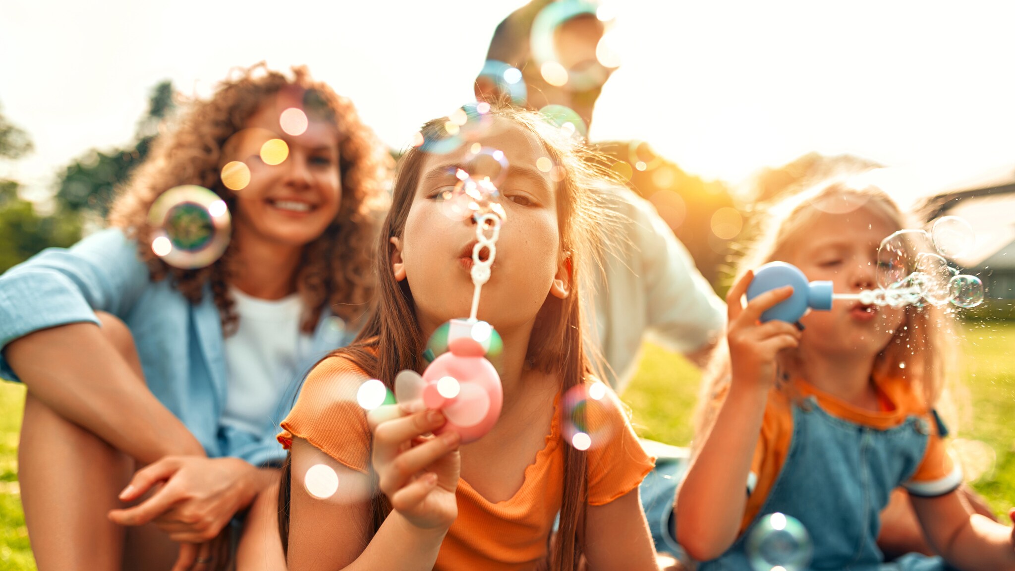Eltern mit Kindern sitzen draußen auf Gras und spielen mit Seifenblasen bei Sonnenlicht.
