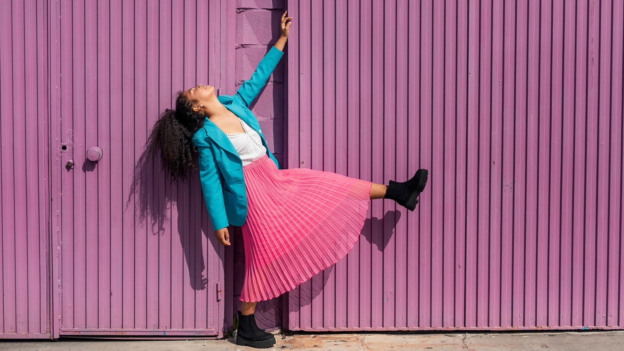 Eine Frau im türkisblauen Balzer und rosafarben Plisseerock posiert vor einer lilafarbenen Fassade. Eine Frau im türkisblauen Balzer und rosafarben Plisseerock posiert vor einer lilafarbenen Fassade.