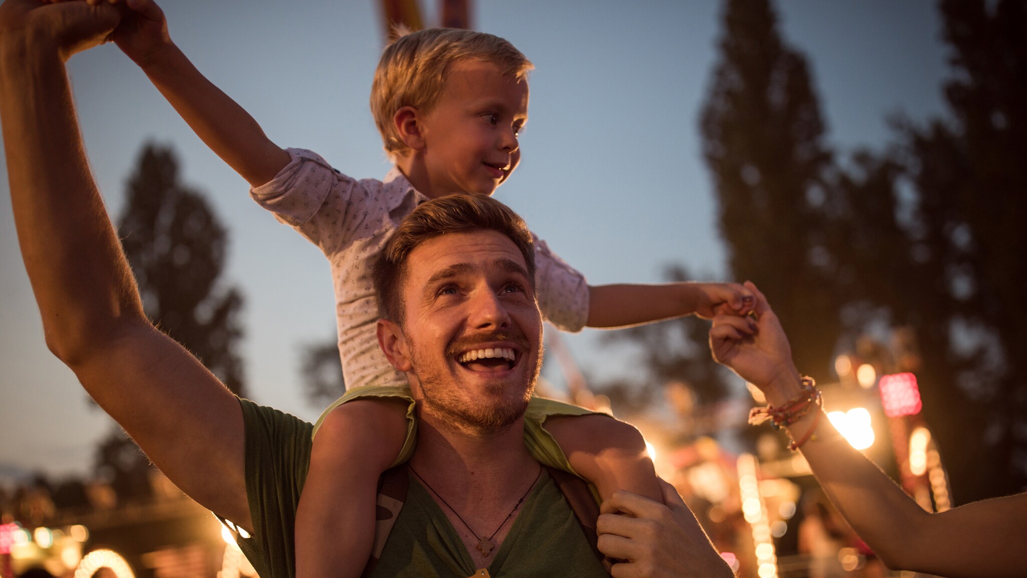 Mann im Dunkeln mit kleinem Kind auf den Schultern, im Hintergrund sind Bäume und Beleuchtung zu sehen.