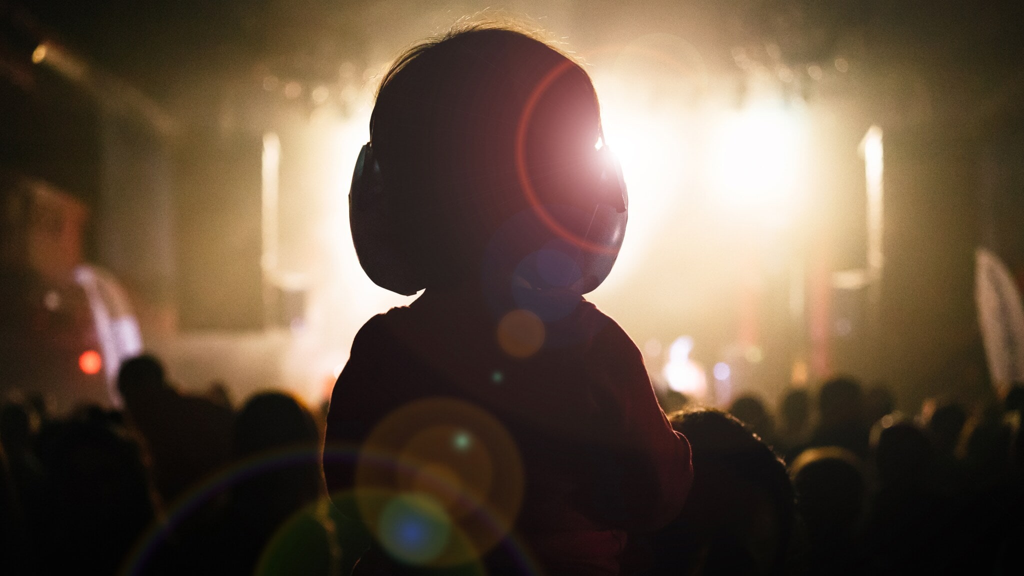 Kind mit Gehörschutz auf den Schultern eines Erwachsenen bei einem Konzert mit Bühnenlicht im Hintergrund. Kind mit Gehörschutz auf den Schultern eines Erwachsenen bei einem Konzert mit Bühnenlicht im Hintergrund.