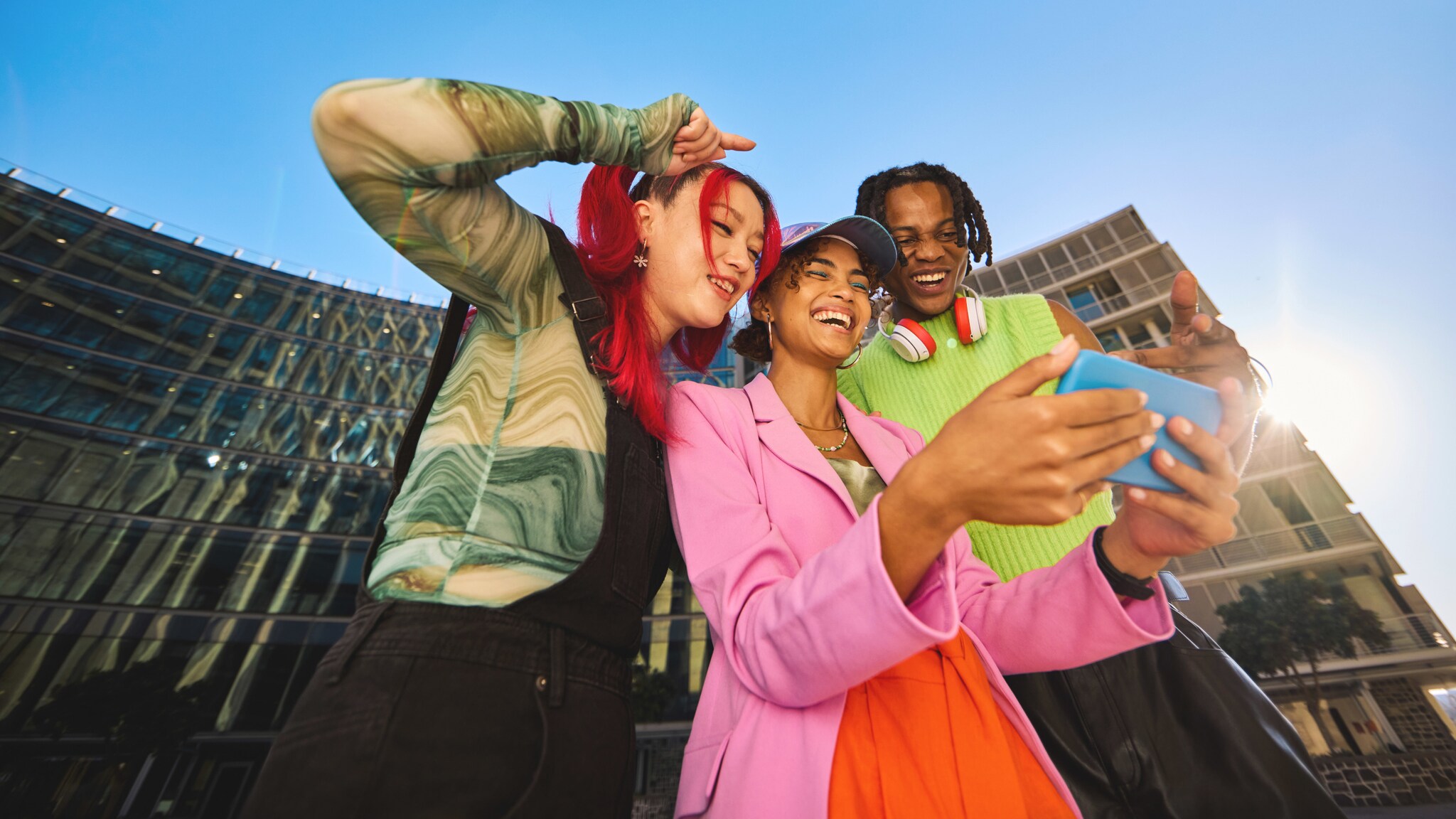 Drei junge, stylische Personen in bunter Kleidung schauen gut gelaunt gemeinsam auf ein Smartphone in urbaner Umgebung unter blauem Himmel.