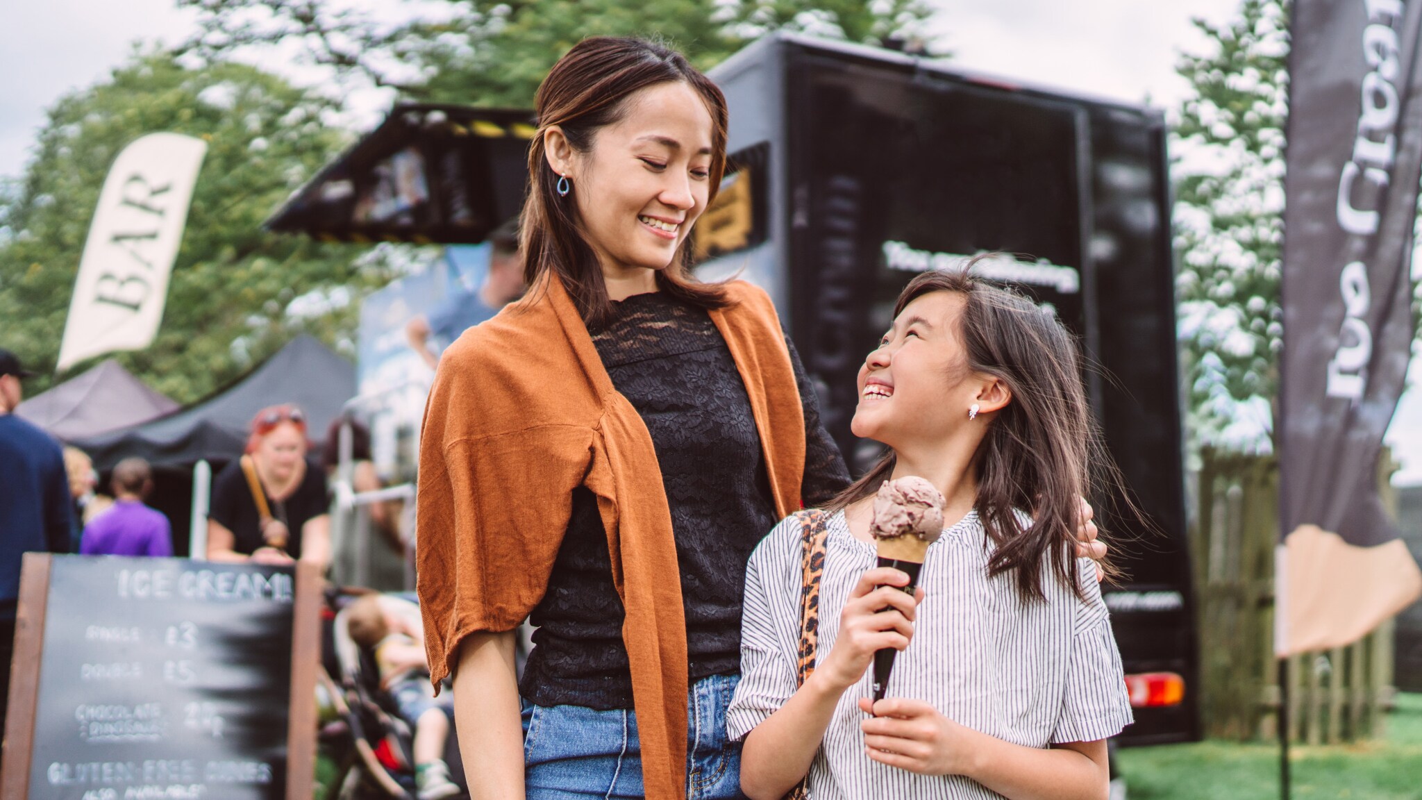Frau mit orangefarbenem Pullover um die Schultern und Kind mit Eiscreme in der Hand auf einem Festival mit Foodtrucks im Hintergrund. Frau mit orangefarbenem Pullover um die Schultern und Kind mit Eiscreme in der Hand auf einem Festival mit Foodtrucks im Hintergrund.
