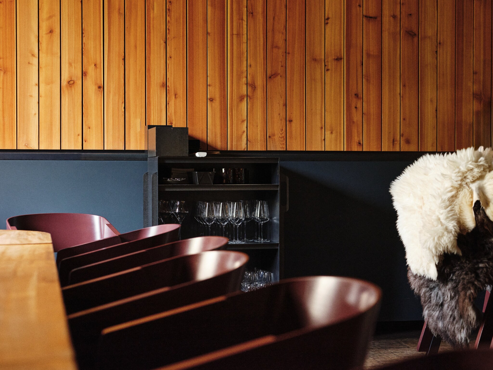 Knapper Ausschnitt des Gastraums mit dunklen Sesseln am langen Tisch und einem kleinen Schrank mit Gläsern, der an der halb holzvertäfelten und halb petrolfarben gestrichenen Wand steht.
