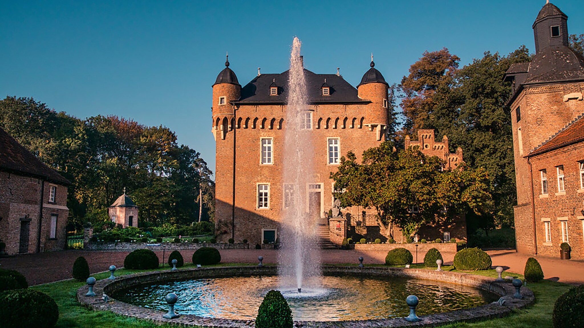 Brunnen mit Fontäne vor einem historischen Schloss mit runden Türmen und Zinnen bei Sonnenuntergang. Brunnen mit Fontäne vor einem historischen Schloss mit runden Türmen und Zinnen bei Sonnenuntergang.