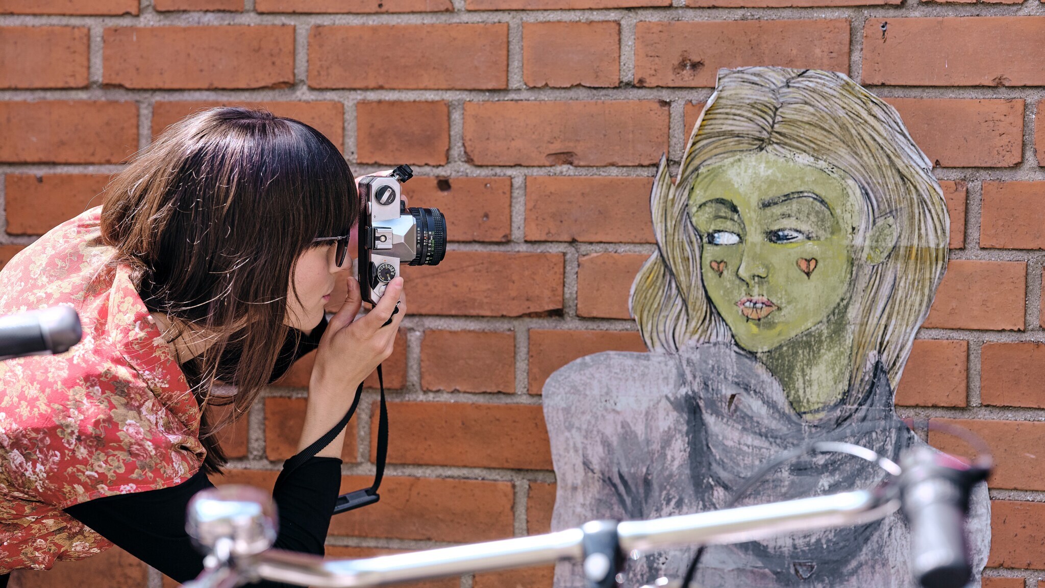 Frau mit braunen Haaren und rotem Blumenmuster-Oberteil fotografiert eine Wandzeichnung einer Person mit langen Haaren vor einer Backsteinmauer.