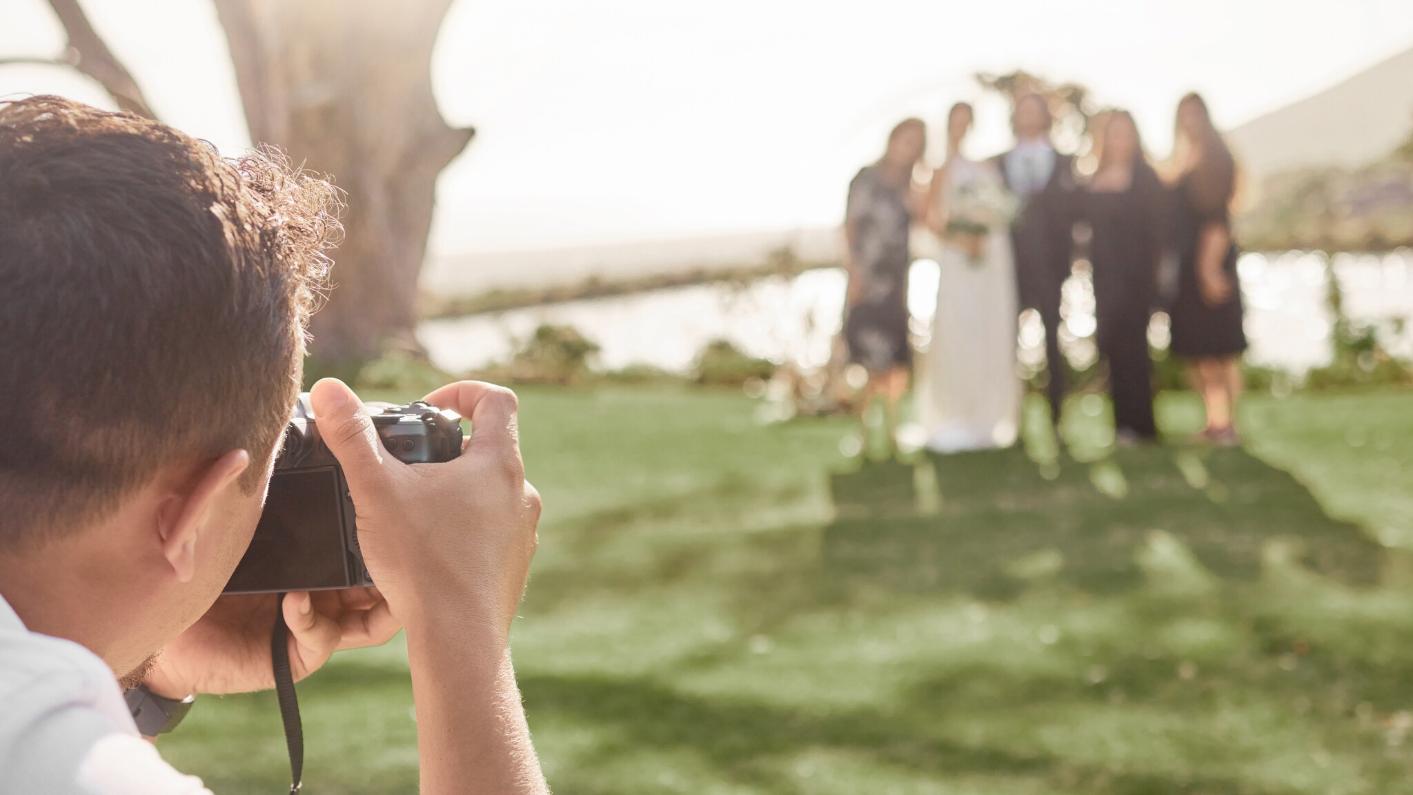 Fotograf fotografiert Brautpaar und Gäste bei Hochzeit im Freien mit See im Hintergrund.