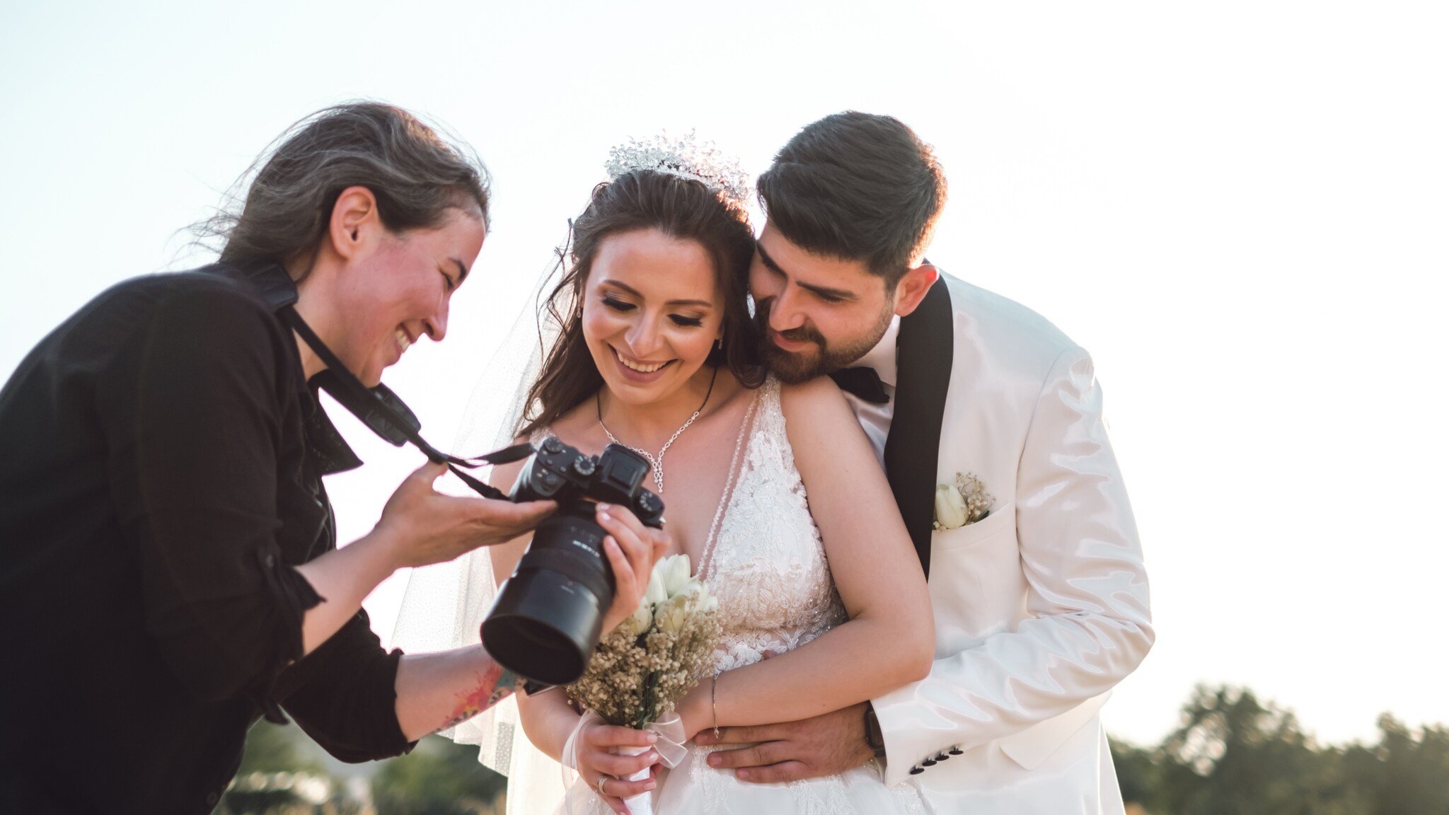 Braut in weißem Spitzenkleid mit Blumenstrauß, Bräutigam in weißem Anzug umarmt sie von hinten, Fotografin zeigt Kamera.