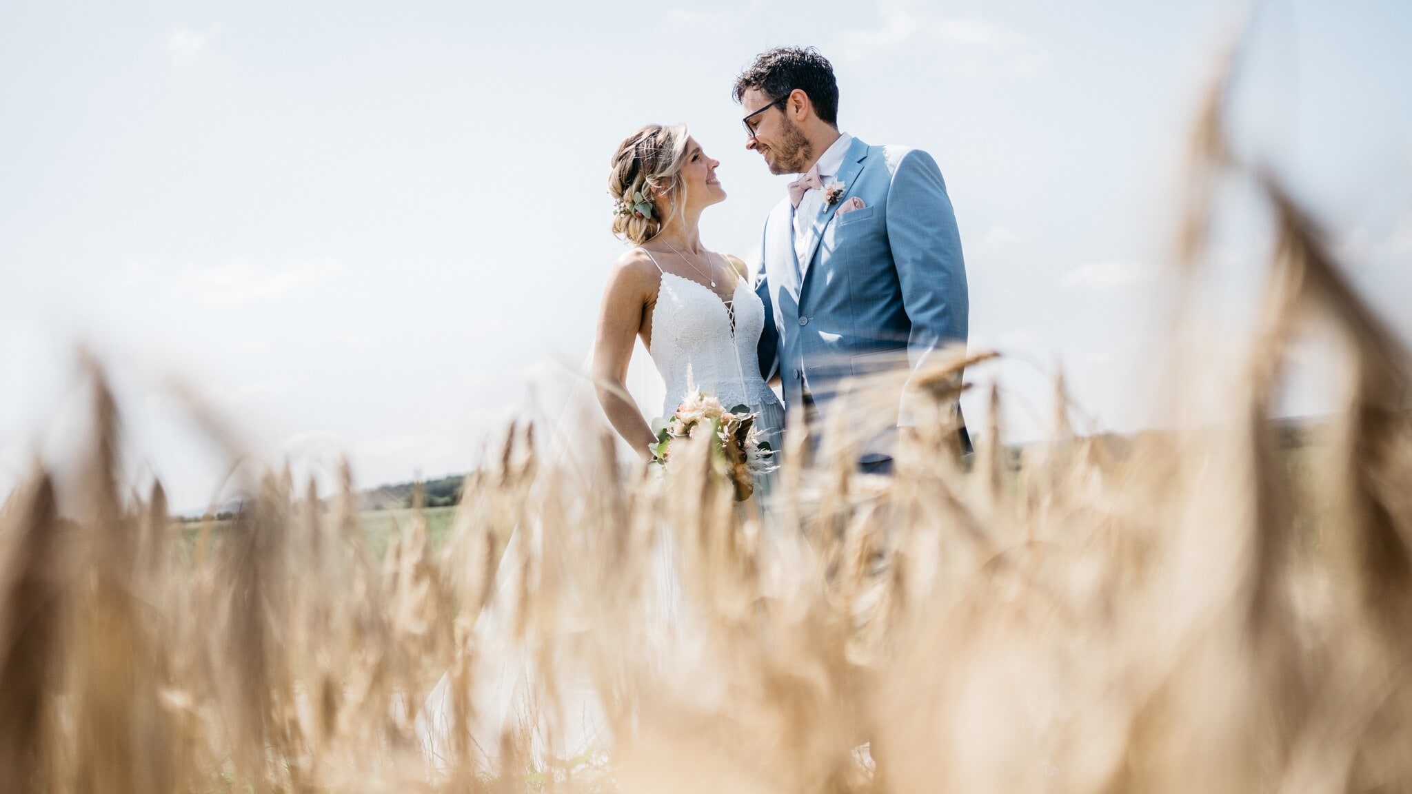 Braut in weißem Kleid mit Blumenstrauß und Bräutigam im blauen Anzug stehen in einem Weizenfeld. Braut in weißem Kleid mit Blumenstrauß und Bräutigam im blauen Anzug stehen in einem Weizenfeld.
