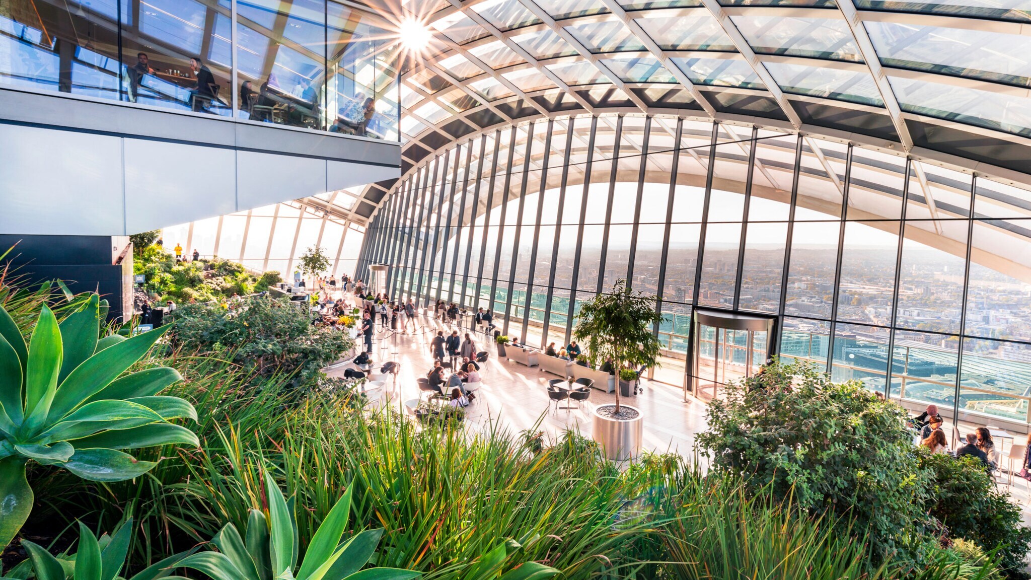 Großer Wintergarten mit Glasdach und Panoramafenstern, viele Pflanzen und Menschen an Tischen, Stadtblick im Hintergrund. Großer Wintergarten mit Glasdach und Panoramafenstern, viele Pflanzen und Menschen an Tischen, Stadtblick im Hintergrund.