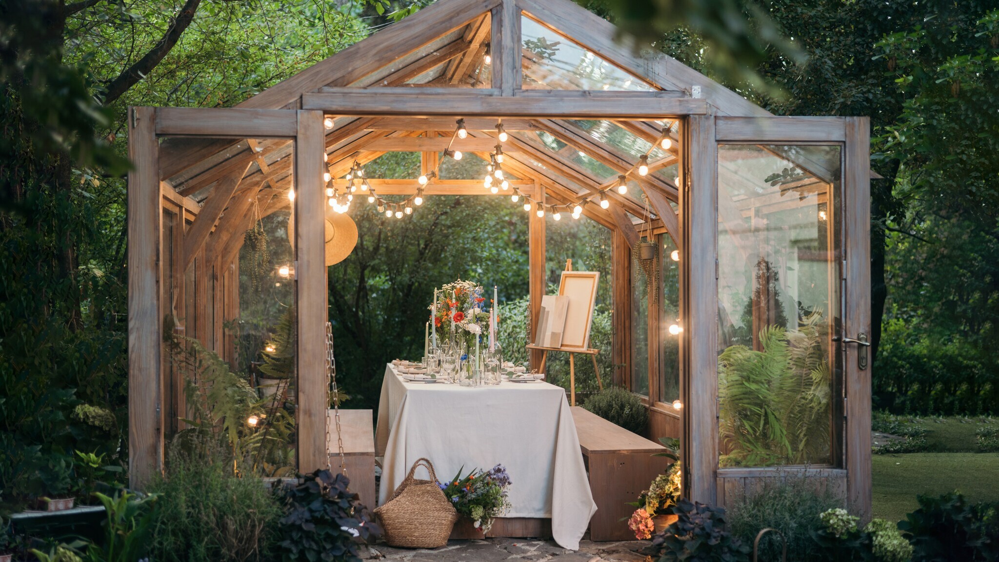 Holz-Gewächshaus mit Glaselementen, innen gedeckter Tisch mit Blumen und Kerzen, Lichterkette am Dach, umgeben von Gartenpflanzen