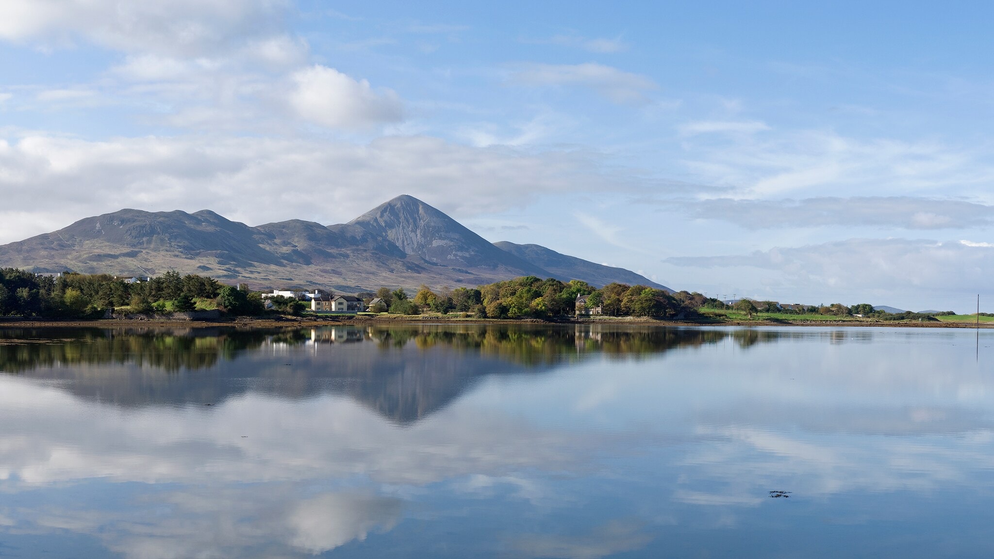 Vor einem großen Gewässer stehen kleine Häuser und Bäume, in Hintergrund ragt ein großer Berg empor, die Szene spiegelt sich in der Wasseroberfläche. Vor einem großen Gewässer stehen kleine Häuser und Bäume, in Hintergrund ragt ein großer Berg empor, die Szene spiegelt sich in der Wasseroberfläche.