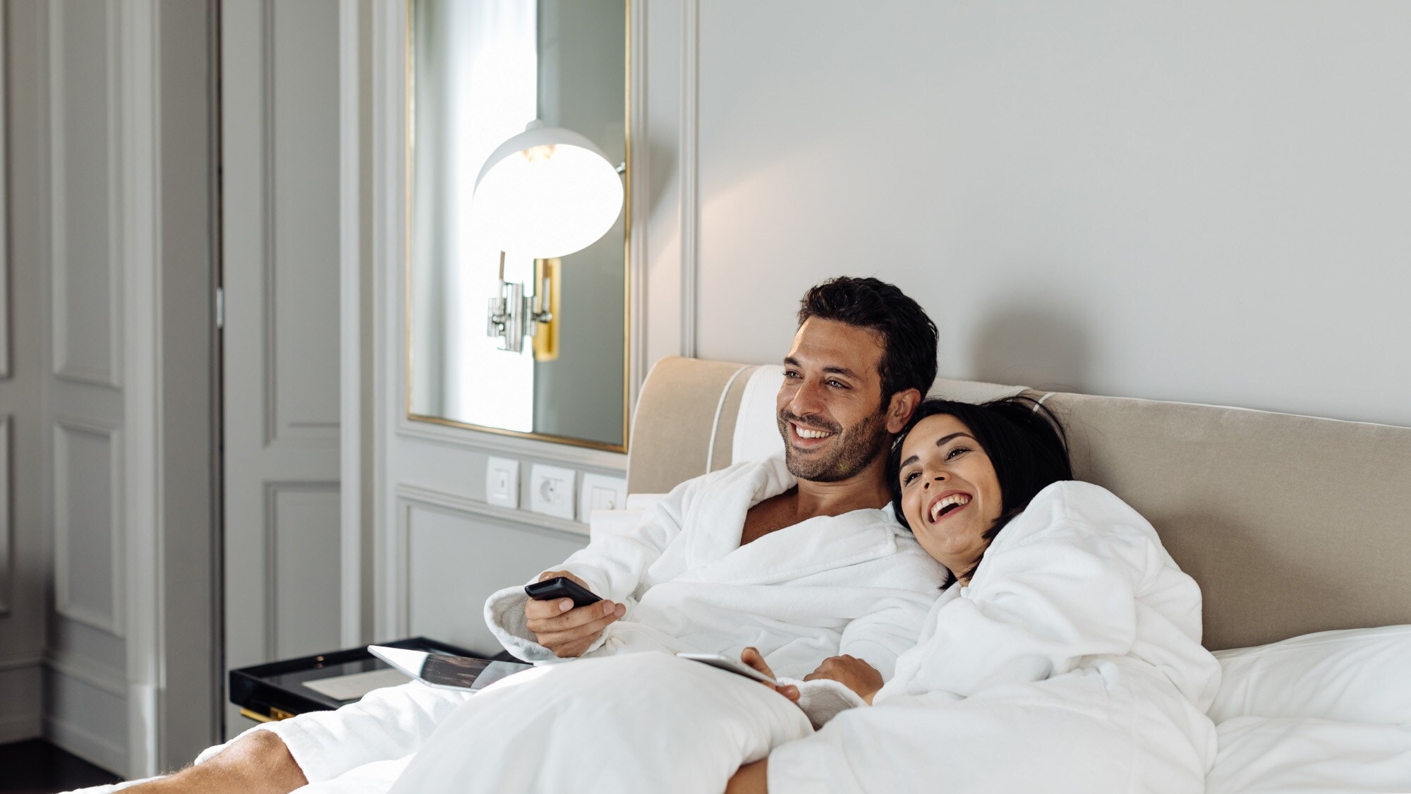 Ein fröhliches Paar in weißen Bademänteln entspannt im Bett einer edlen Hotelsuite Ein fröhliches Paar in weißen Bademänteln entspannt im Bett einer edlen Hotelsuite