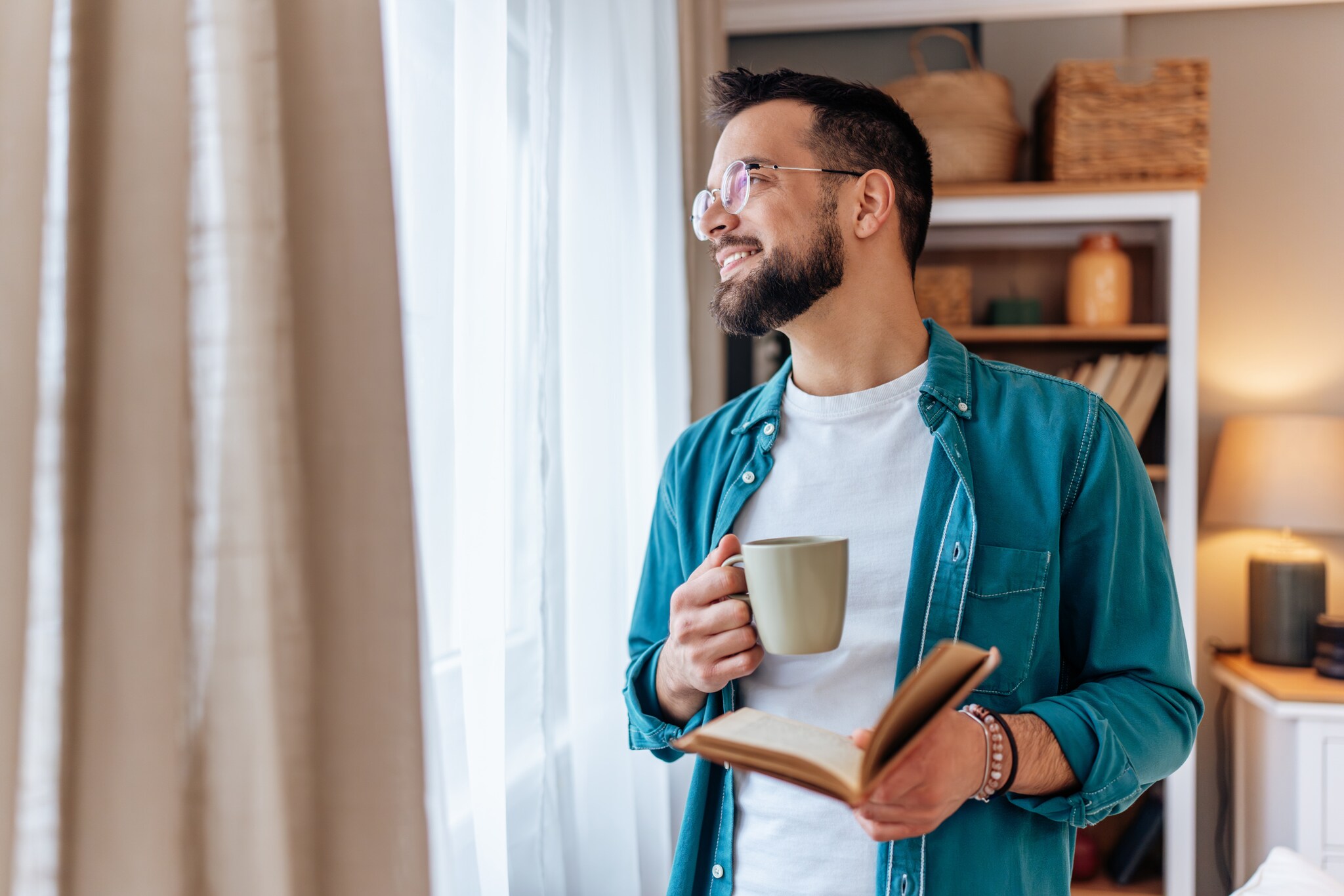 Ein Mann im Jeanshemd mit Buch und Kaffeebecher in den Händen schaut entspannt aus einem Fenster mit Vorhängen. Ein Mann im Jeanshemd mit Buch und Kaffeebecher in den Händen schaut entspannt aus einem Fenster mit Vorhängen.