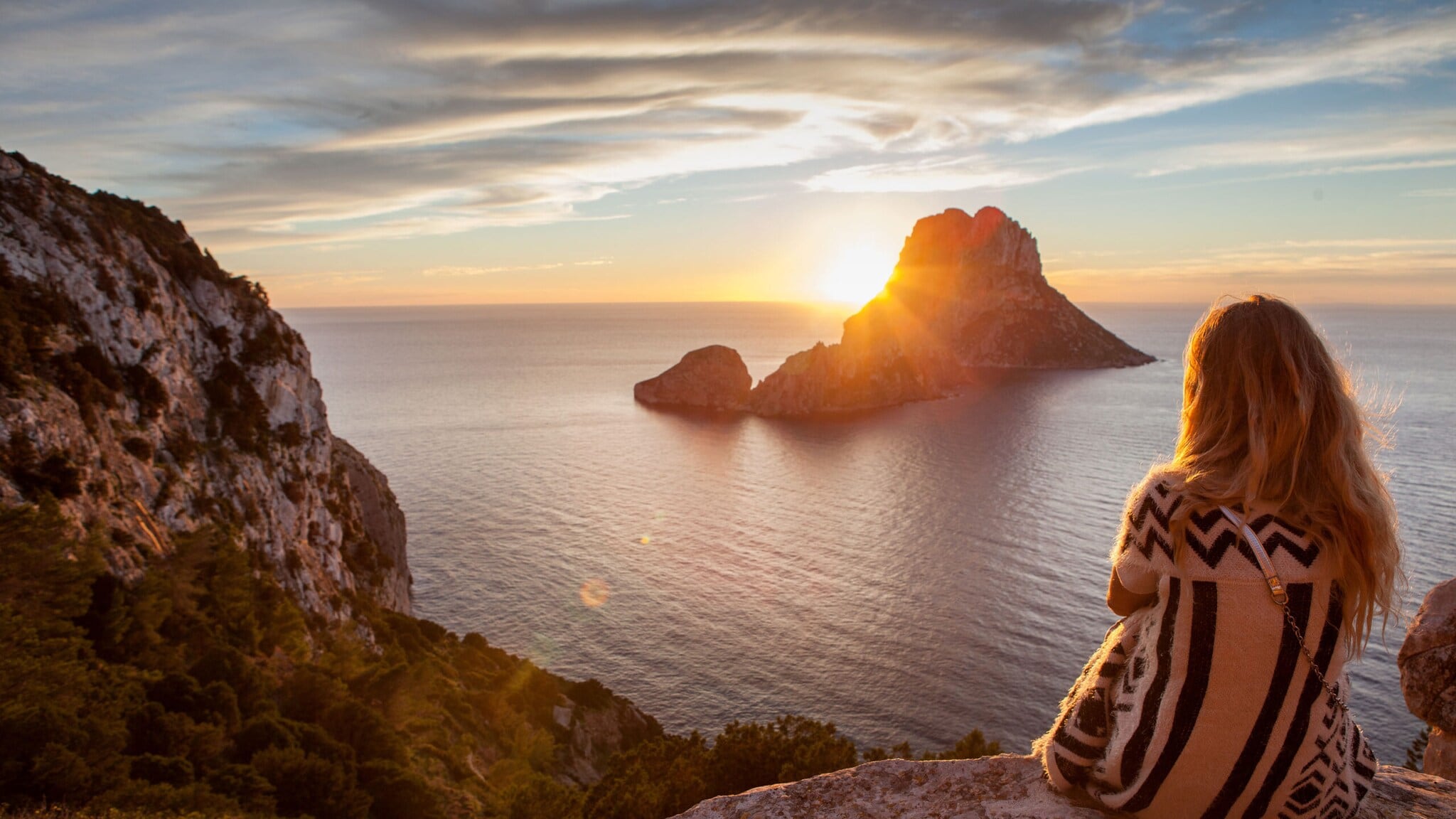 Frau mit langem Haar und gestreiftem Pullover sitzt auf Felsen und blickt auf Sonnenuntergang über Meer und Insel Frau mit langem Haar und gestreiftem Pullover sitzt auf Felsen und blickt auf Sonnenuntergang über Meer und Insel