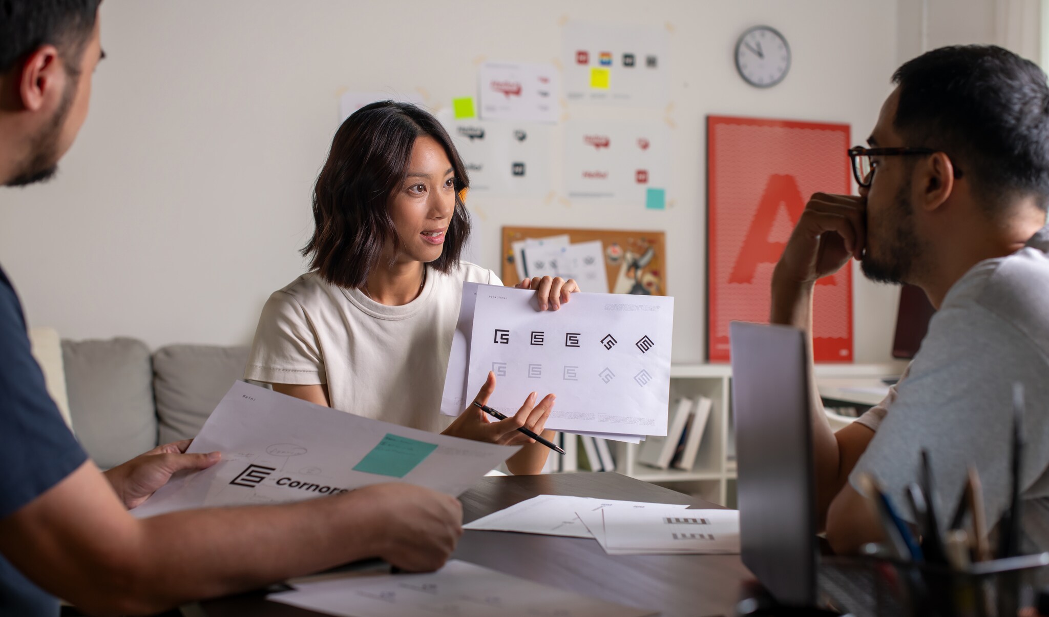 Eine Frau sitzt an einem Tisch zwischen zwei weiteren Personen und präsentiert Logoentwürfe auf einem Papierblatt in ihren Händen in einem Arbeitszimmer.