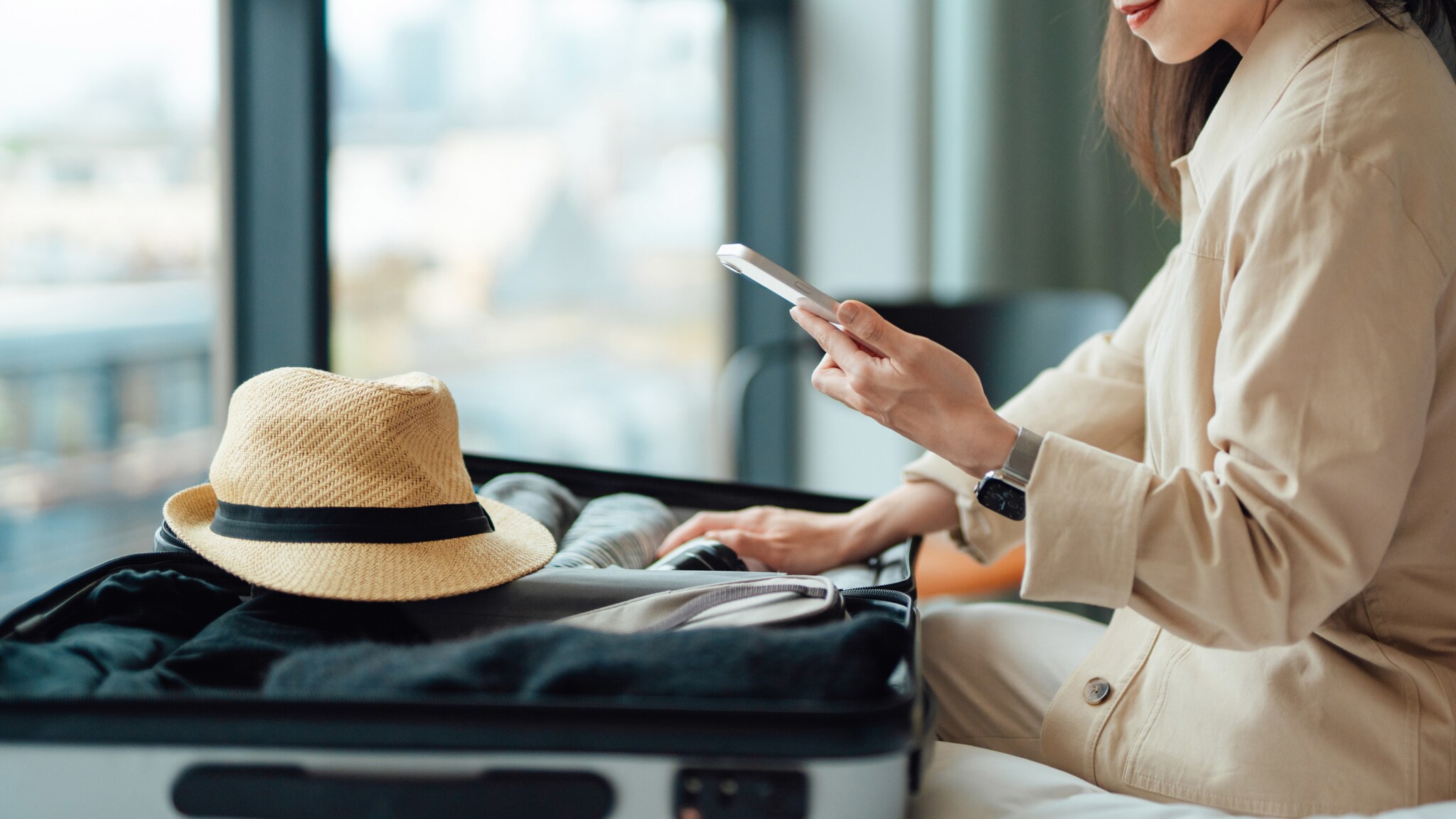 Eine Frau hält ein Smartphone über einen offenen, gepackten Trolley mit Sonnenhut auf einem Bett.