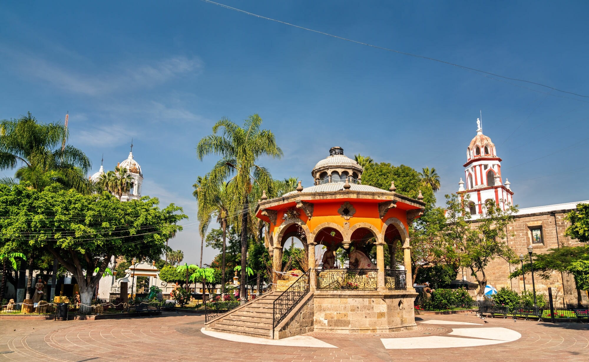 Ein Kiosk auf einem Platz mit viel Grün, im Hintergrund der Turm einer weiß-roten Kirche und blauer Himmel. Ein Kiosk auf einem Platz mit viel Grün, im Hintergrund der Turm einer weiß-roten Kirche und blauer Himmel.