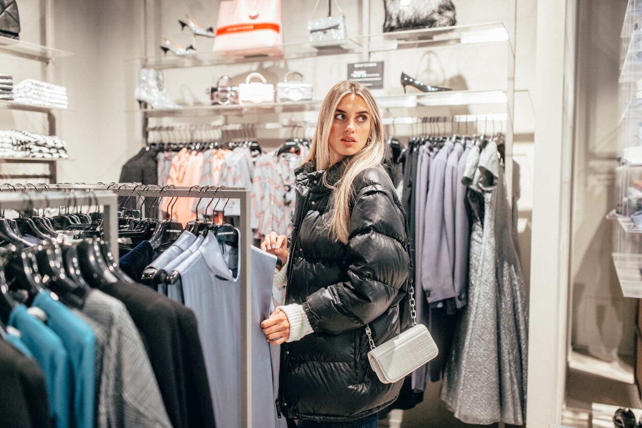 Eine Frau in schwarzer Pufferjacke zwischen Shoppingständern, auf denen Damenbekleidung hängt