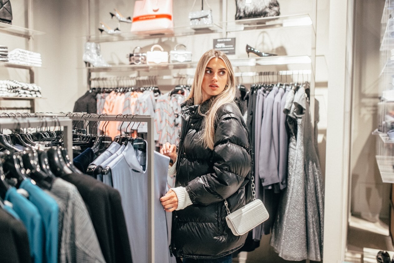 Eine Frau in schwarzer Pufferjacke zwischen Shoppingständern, auf denen Damenbekleidung hängt Eine Frau in schwarzer Pufferjacke zwischen Shoppingständern, auf denen Damenbekleidung hängt