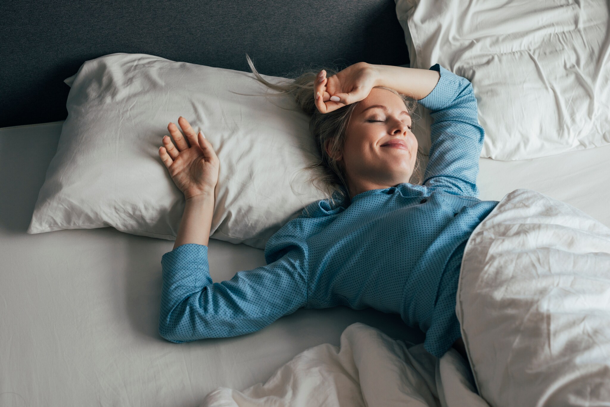 Eine zufrieden lächelnde Frau im blauen Pyjama liegt mit geschlossenen Augen in einem Bett mit weißer Bettwäsche bei Tageslicht. Eine zufrieden lächelnde Frau im blauen Pyjama liegt mit geschlossenen Augen in einem Bett mit weißer Bettwäsche bei Tageslicht.