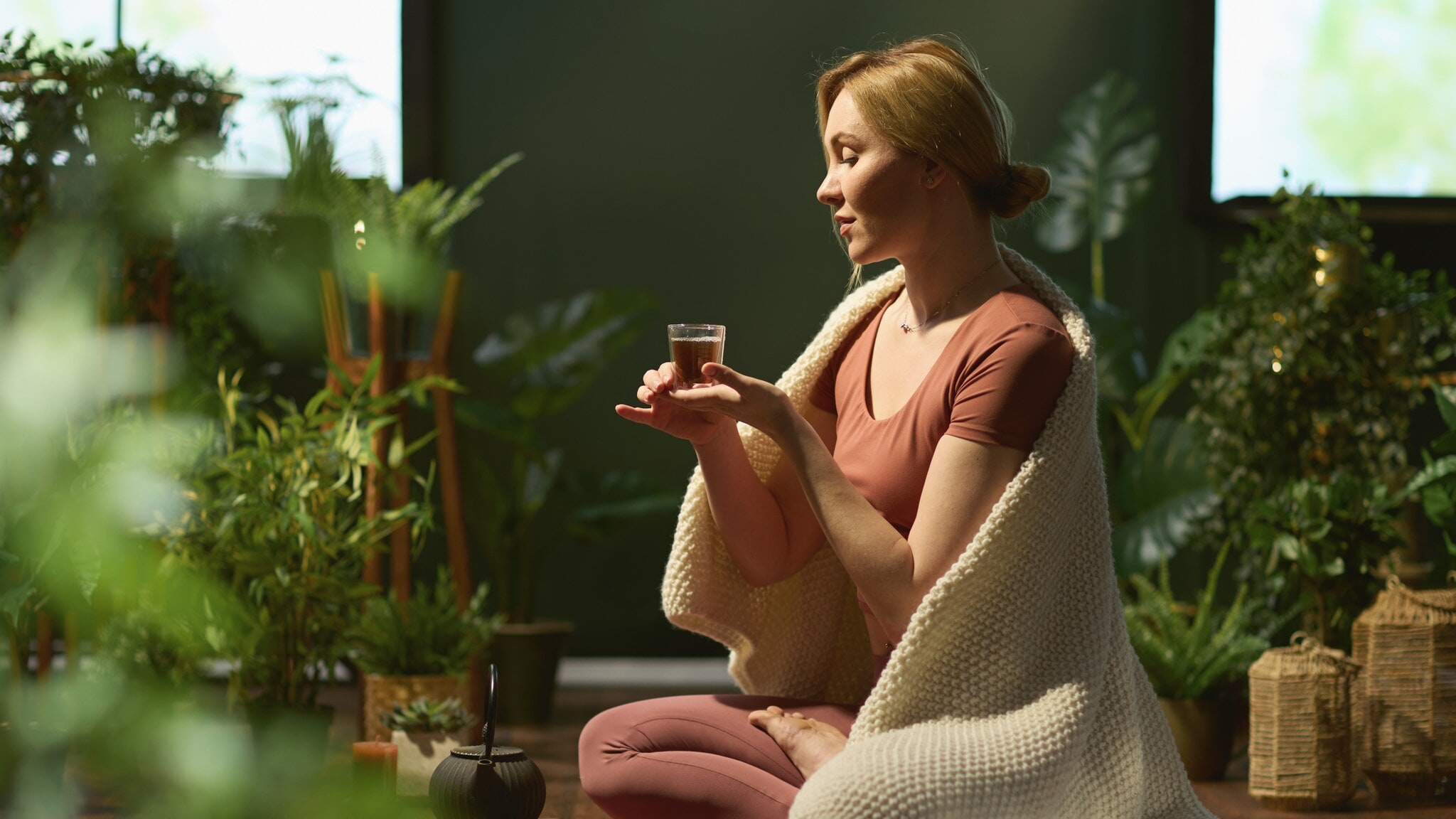 Frau sitzt im Schneidersitz auf dem Boden, hält eine kleine Tasse, trägt rosa Sportkleidung und eine weiße Decke über den Schultern, umgeben von Pflanzen. Frau sitzt im Schneidersitz auf dem Boden, hält eine kleine Tasse, trägt rosa Sportkleidung und eine weiße Decke über den Schultern, umgeben von Pflanzen.