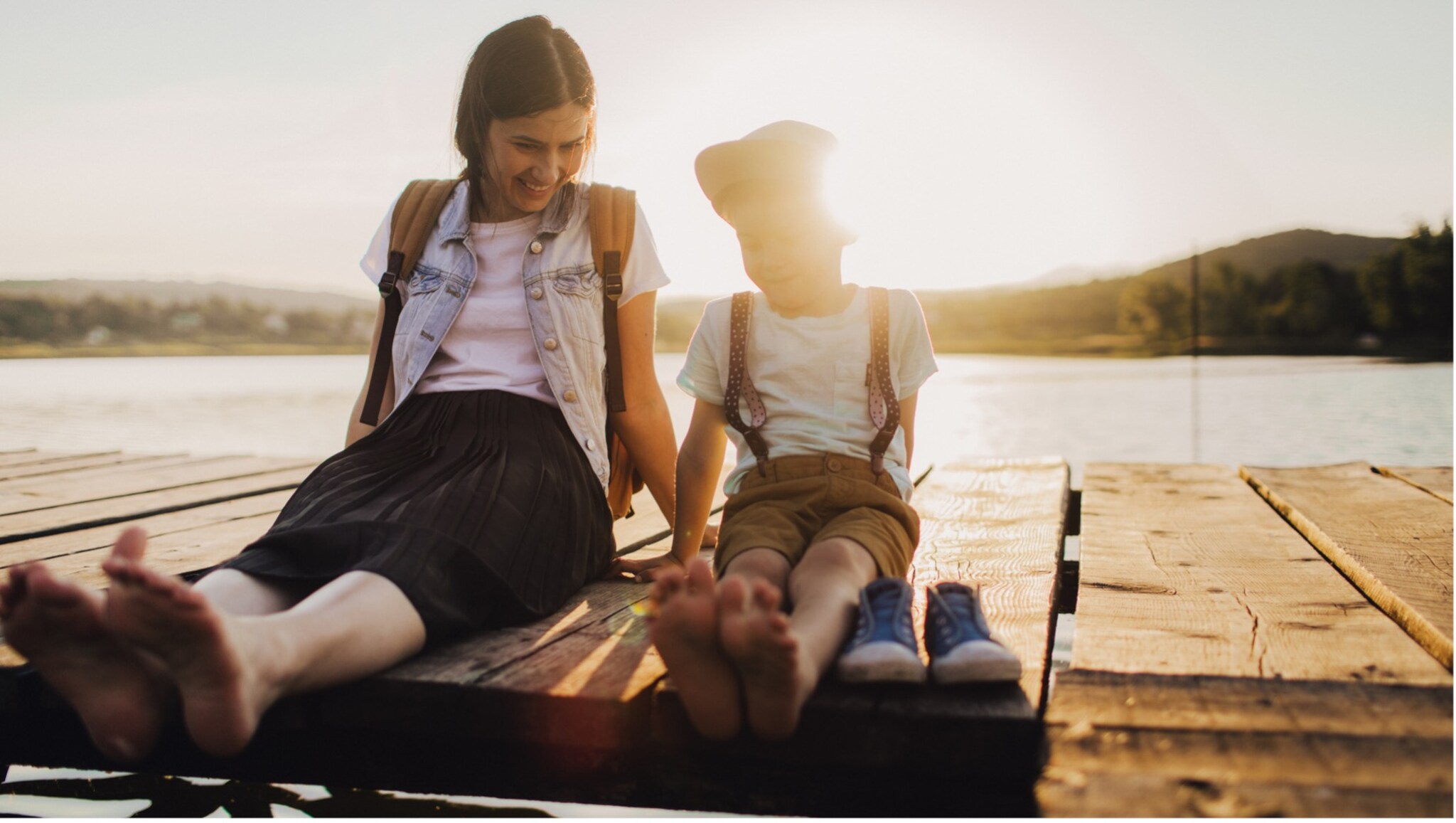 Mutter und Kind sitzen barfuß auf einem Holzsteg am See, die Sonne scheint im Hintergrund