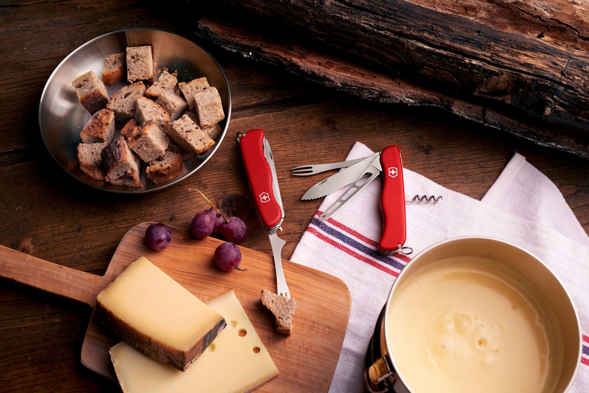 Zwei aufgeklappte, rote Schweizer Taschenmesser mit Käsemesser und Fonduegabel auf einem Holztisch neben Käsescheiben und einem Fonduetopf