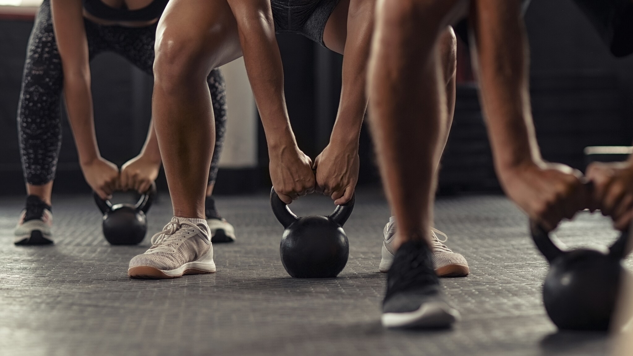 Drei Personen in Sportkleidung halten Kettlebells mit beiden Händen, Blick auf die Beine und Hände, Fitnessstudio-Boden sichtbar.